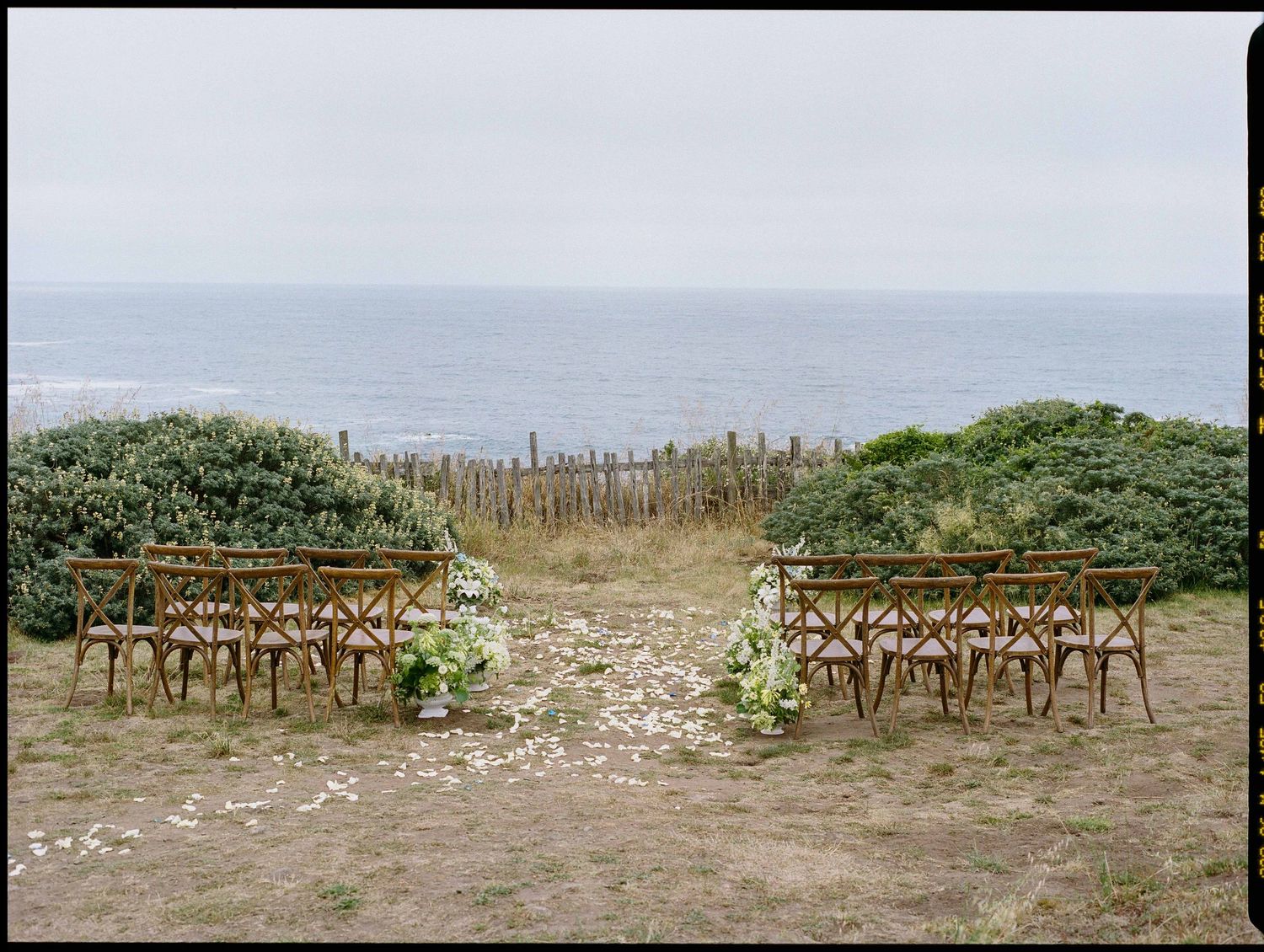 Outdoor coastal wedding ceremony setup with wooden chairs and floral arrangements overlooking the ocean.