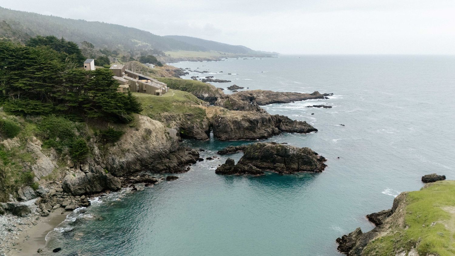 Aerial view of rocky California coastline with turquoise waters, cliffs, and white buildings nestled among evergreen trees.