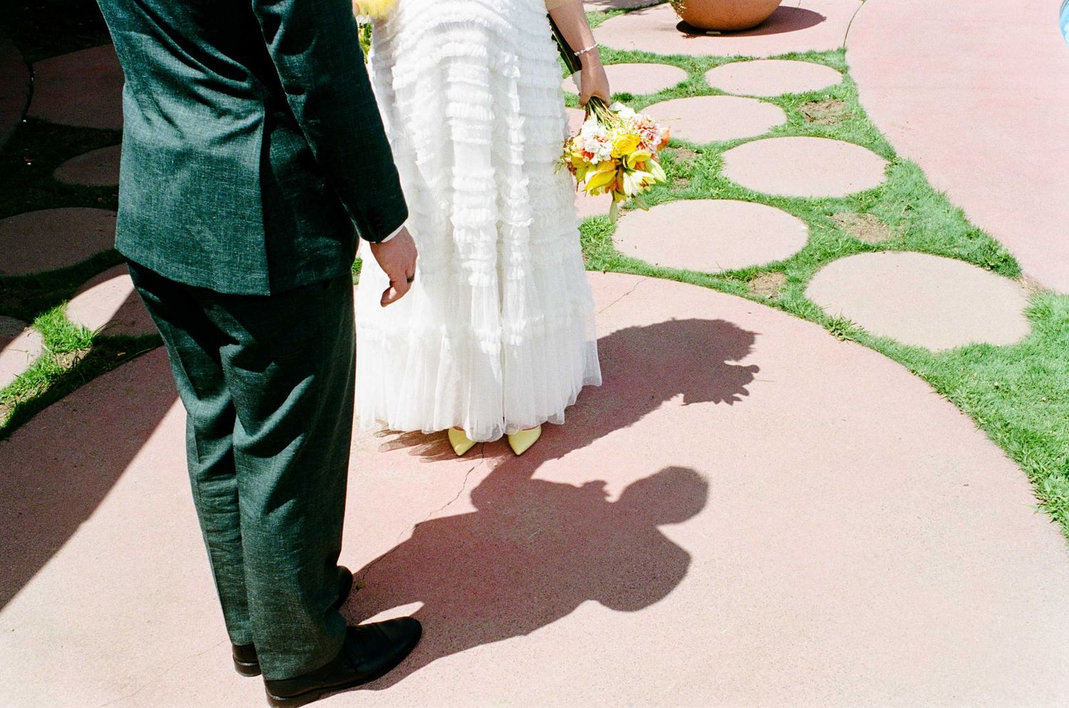 Bride and groom walking hand in hand, casting shadows on a pink pathway, bride holding yellow bouquet.