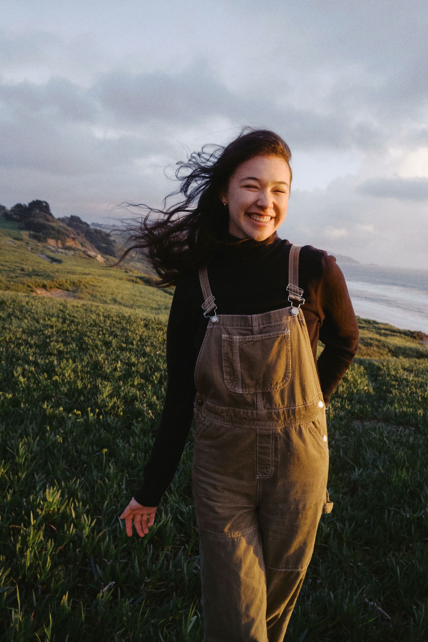 Smiling young woman in overalls and black turtleneck standing on coastal hillside with wind blowing her hair.