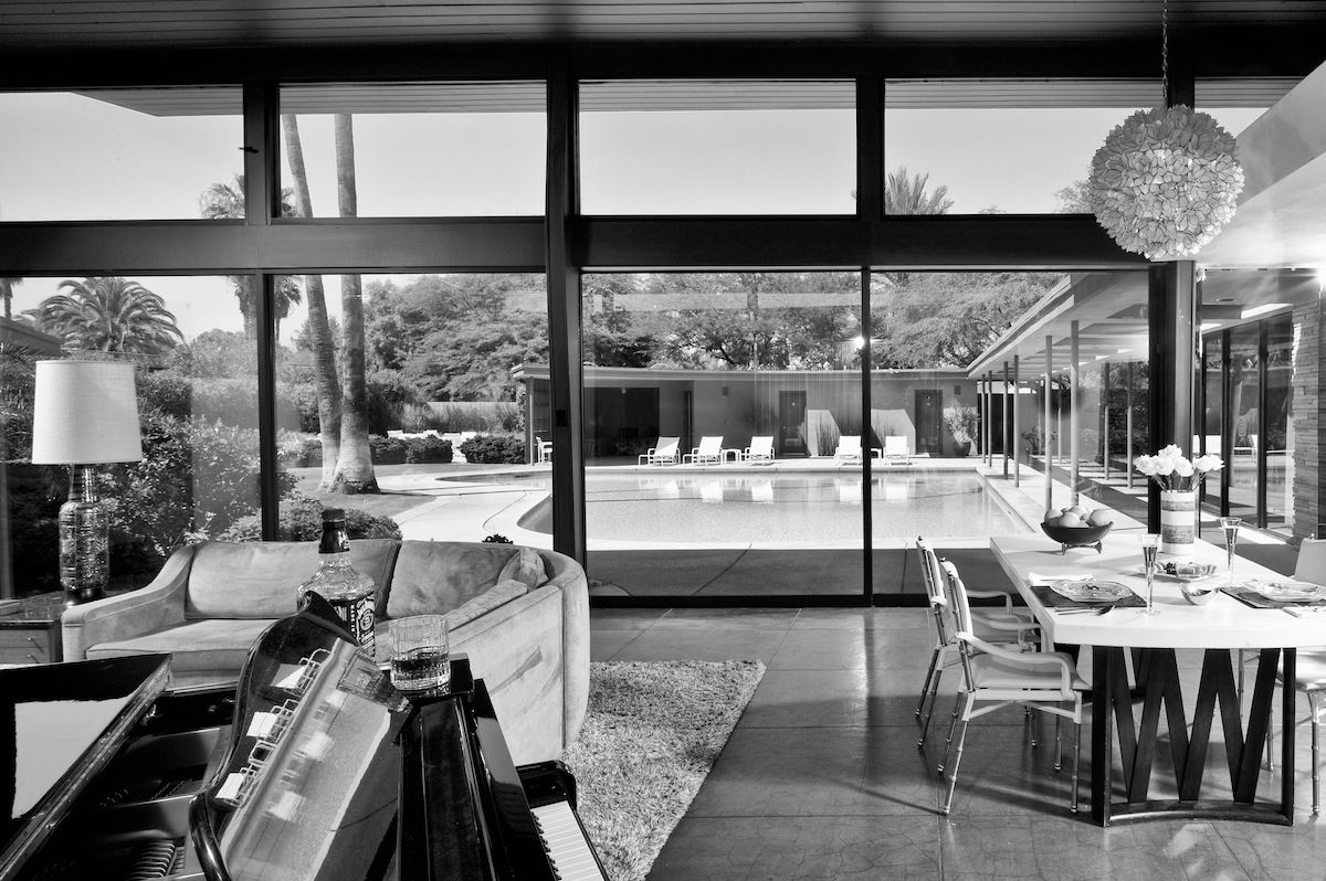 Black and white photo of a mid-century modern interior with glass walls overlooking a pool and palm trees.