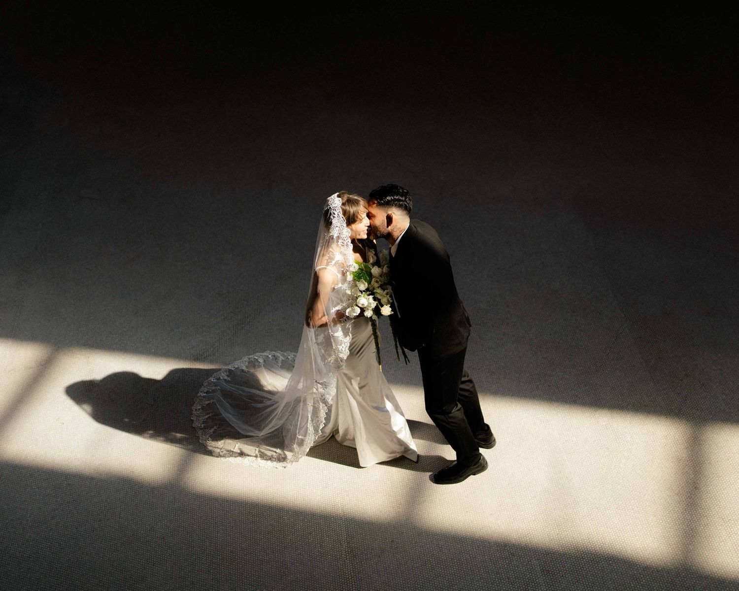 Aerial view of bride and groom sharing a kiss, bride in white gown with veil holding bouquet, groom in black suit.
