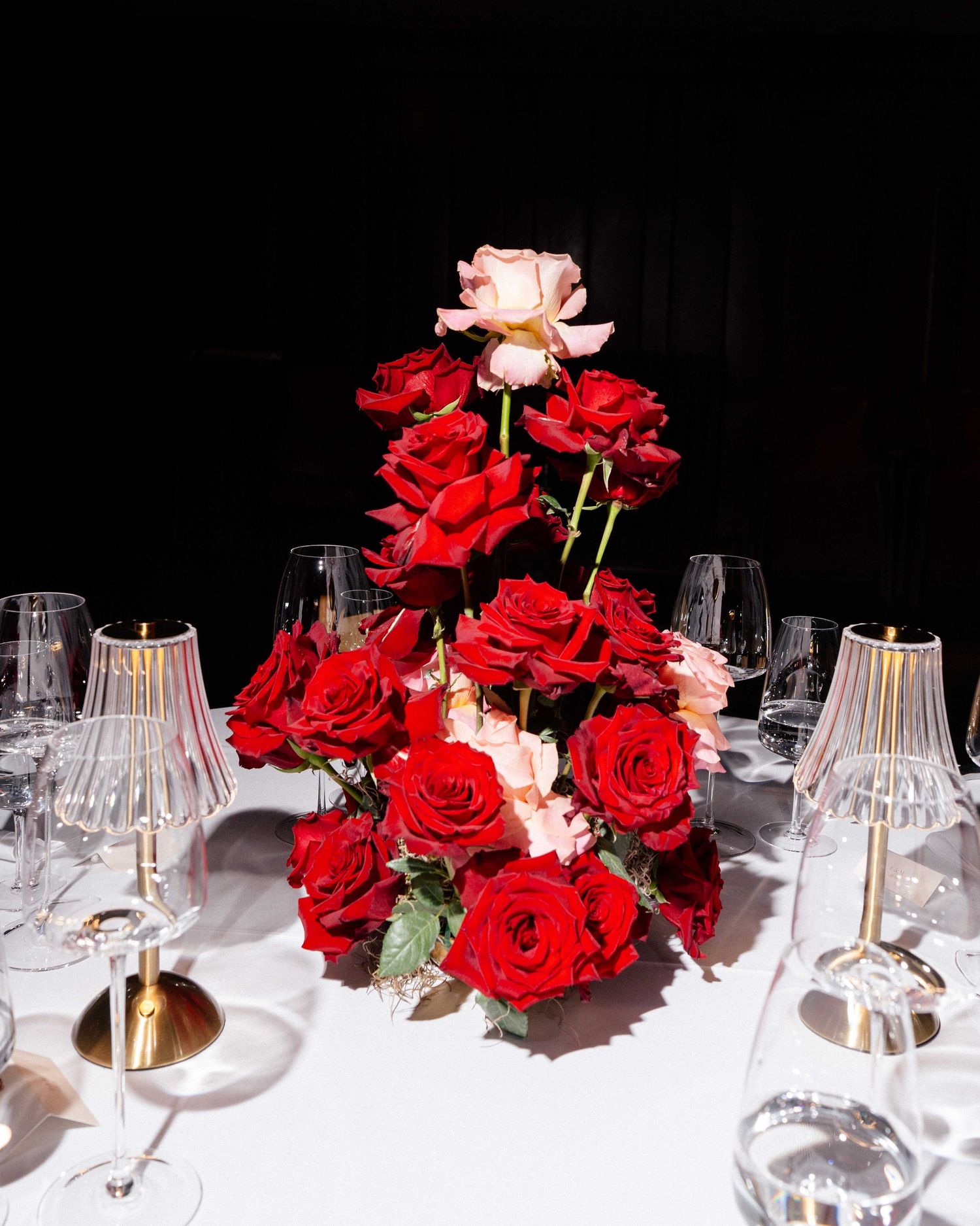 Elegant red and pink rose centerpiece arranged in a tiered display on a white tablecloth with crystal glasses and gold candlesticks.
