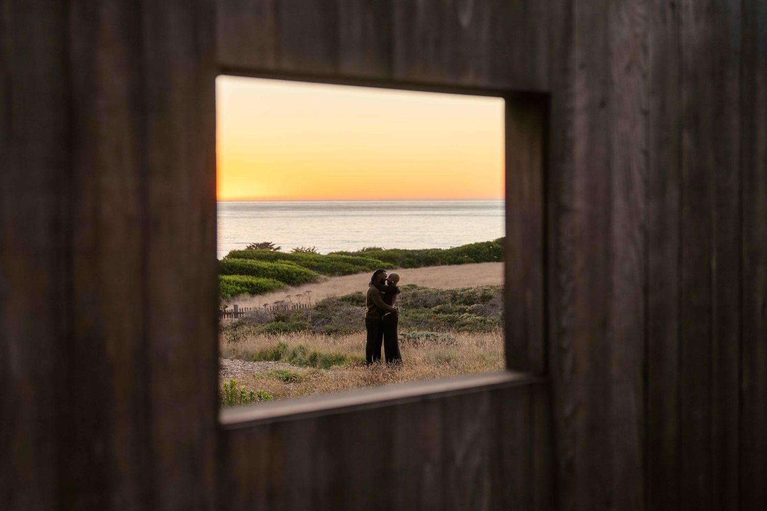 Couple embracing at sunset framed by a rustic wooden window overlooking a coastal landscape with golden sky.