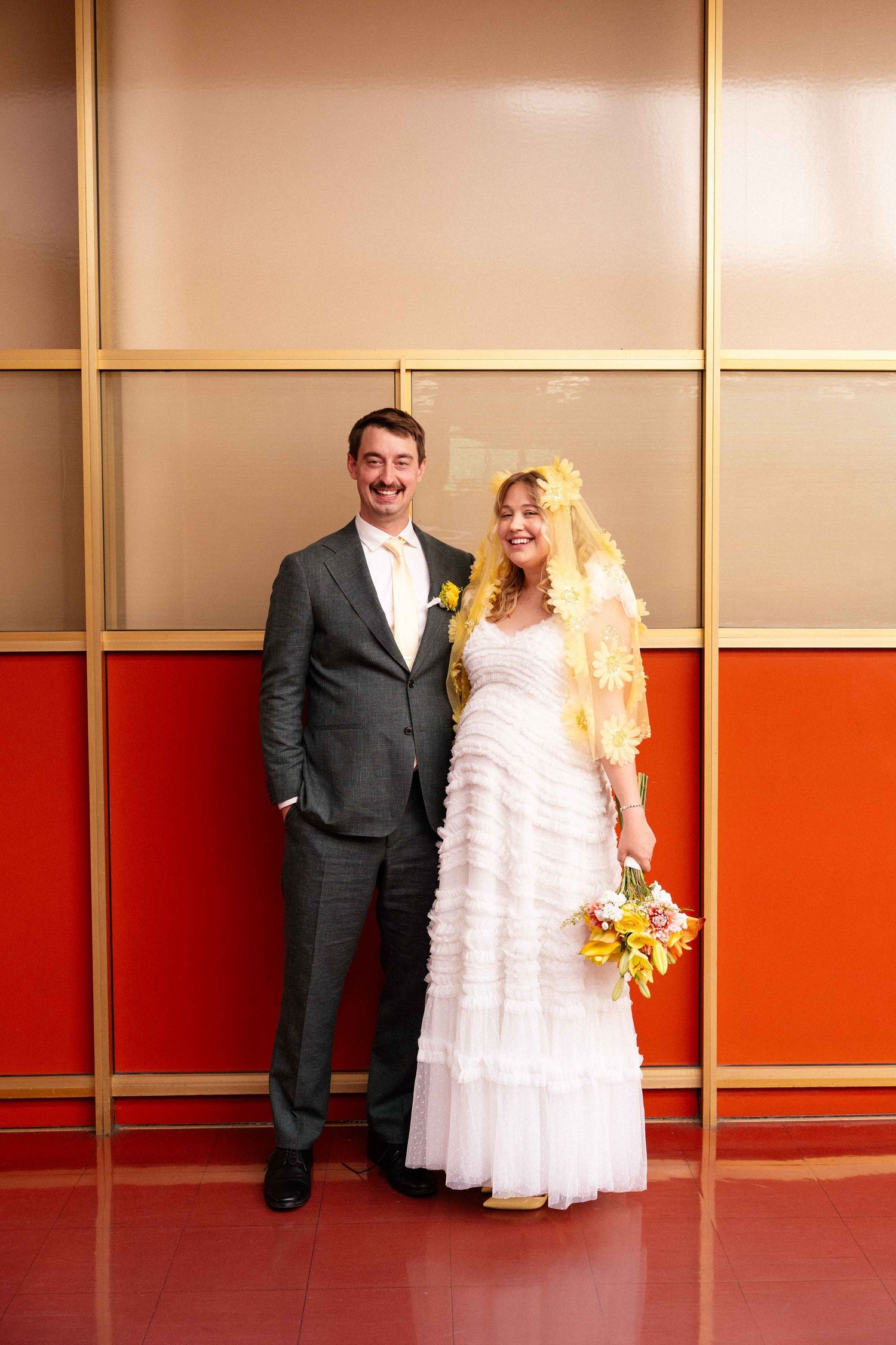 Bride in white ruffled gown with yellow veil and groom in gray suit pose together at their wedding.