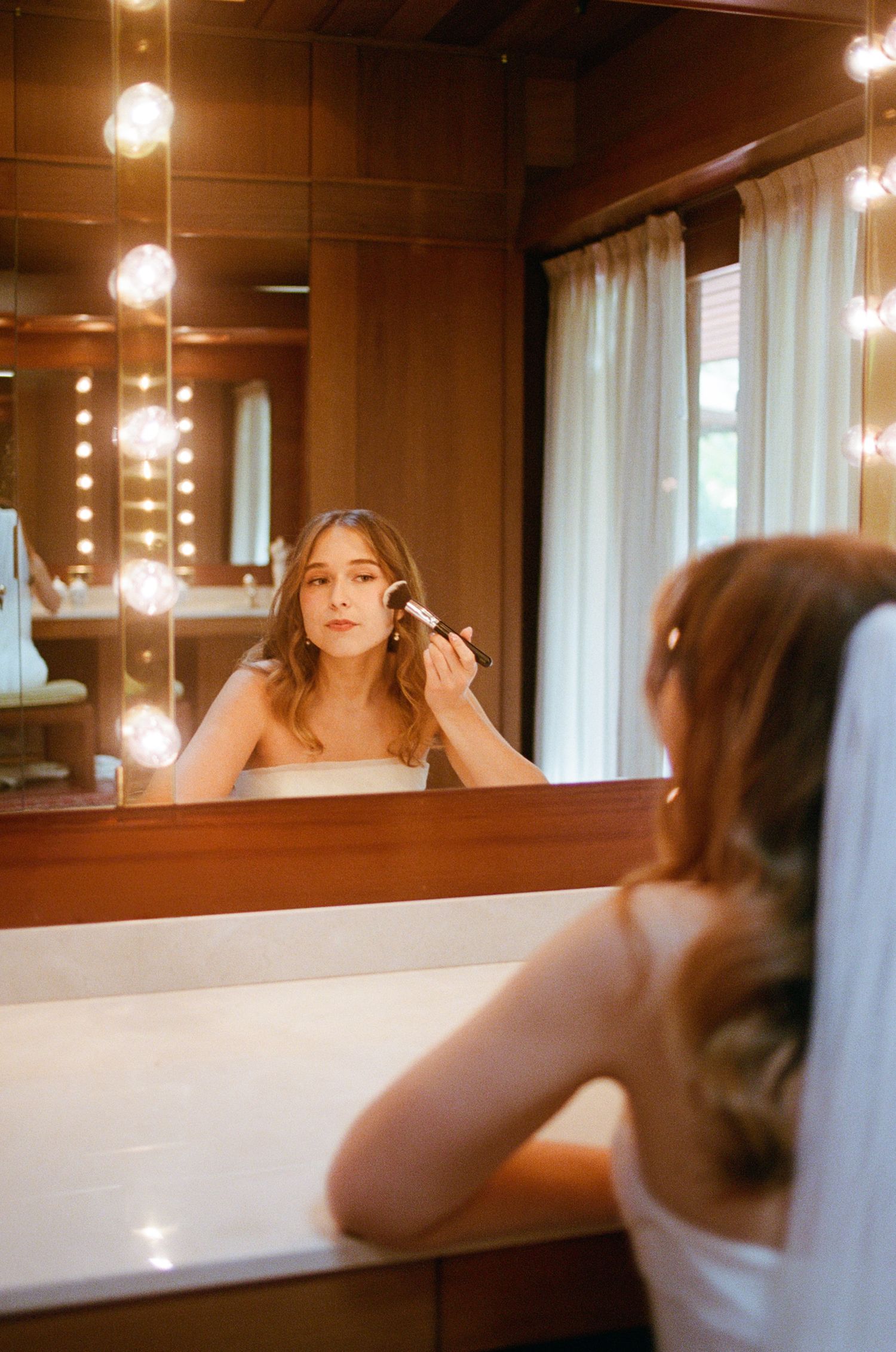 Bride applying blush makeup at a Hollywood vanity mirror with warm glowing bulb lights in a wood-paneled bathroom.