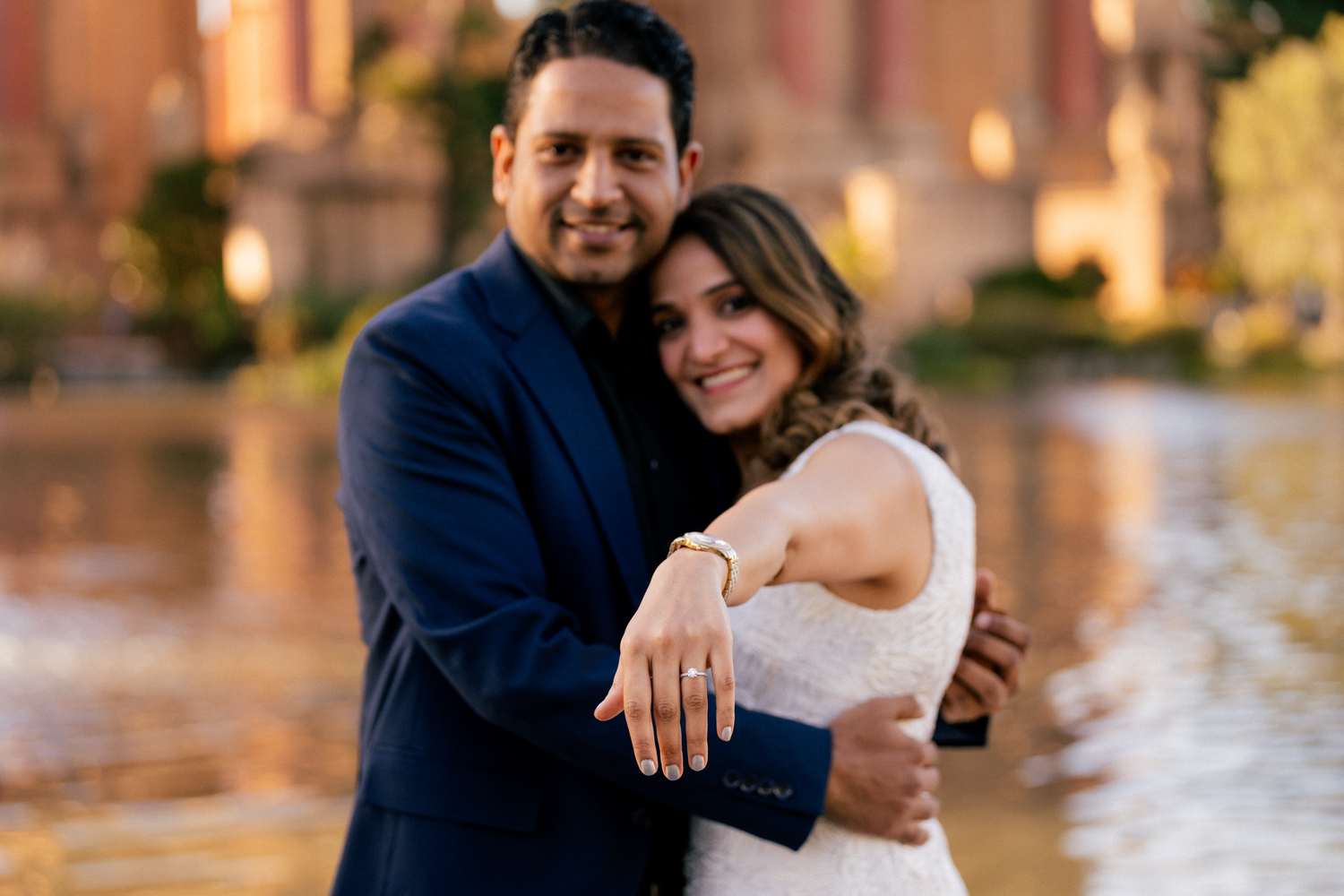 A romantic engagement portrait with a couple embracing near water at sunset.