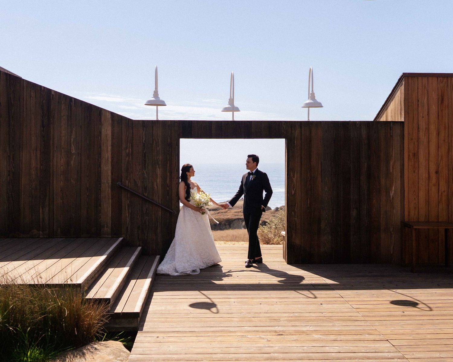 Bride and groom holding hands on a wooden deck at a coastal venue with ocean views on their wedding day.