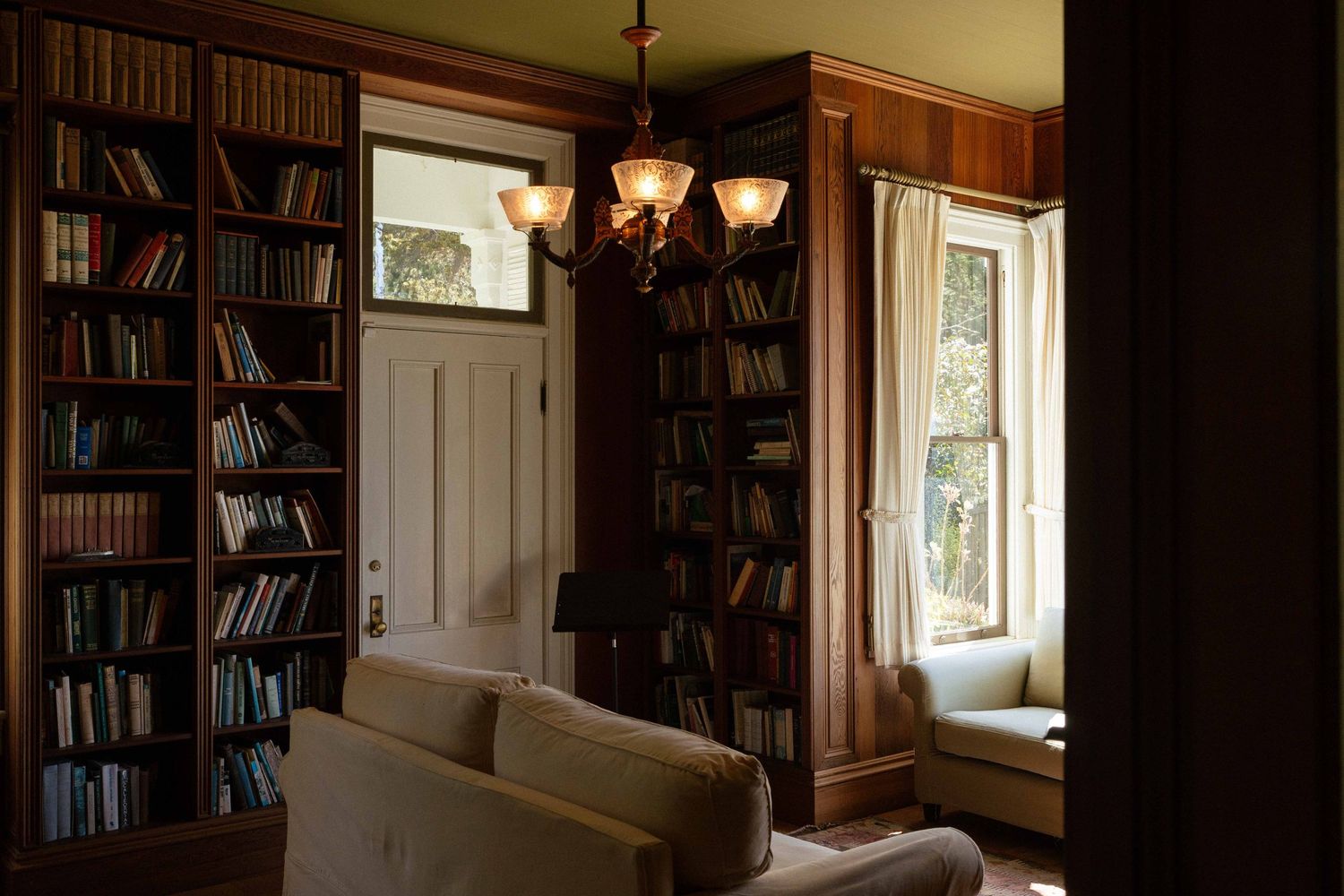 Cozy home library corner with built-in wooden bookshelves, antique chandelier, and soft natural light from window.
