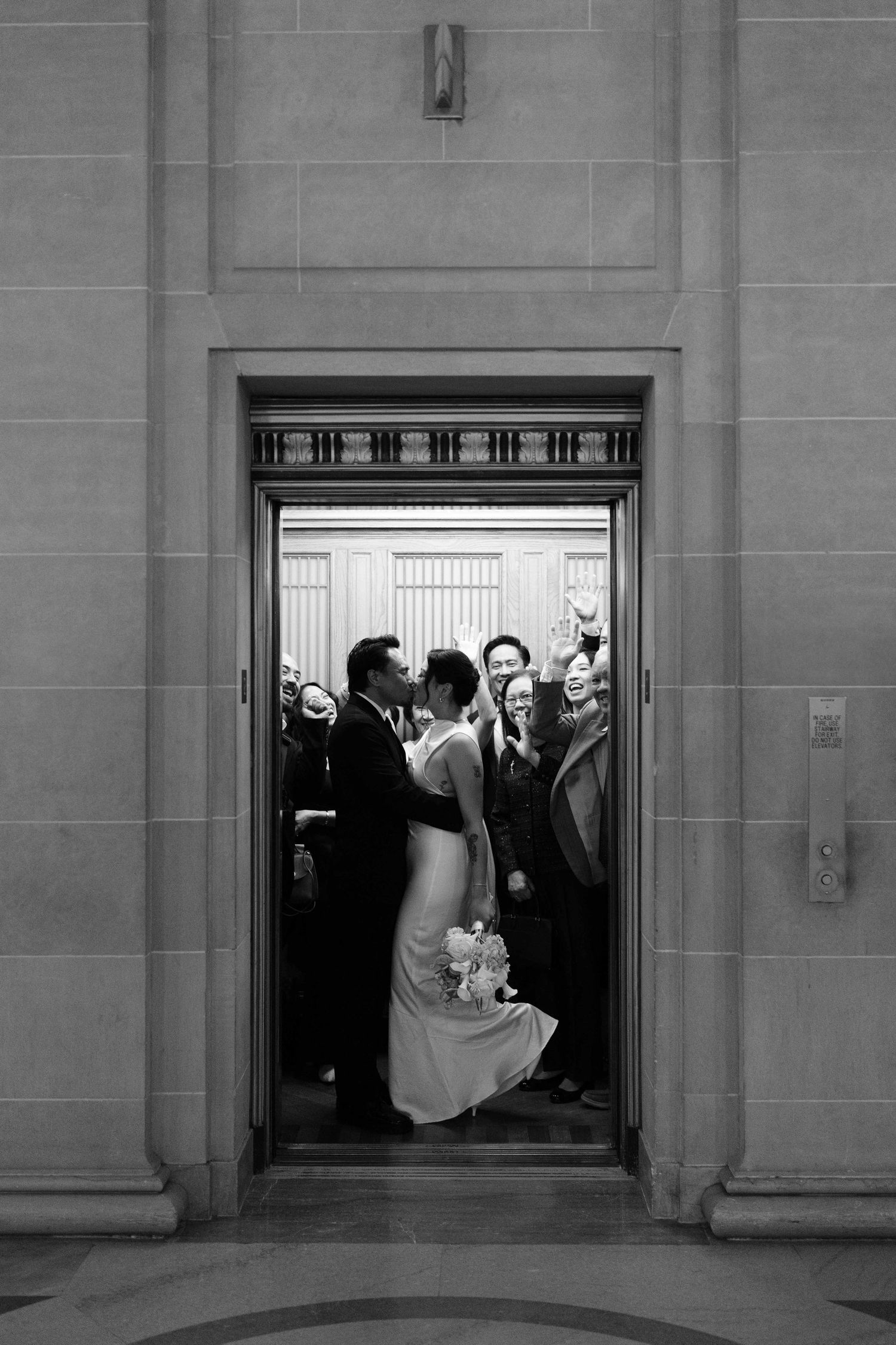 Black and white photo of bride and groom kissing in elevator doorway surrounded by cheering guests.