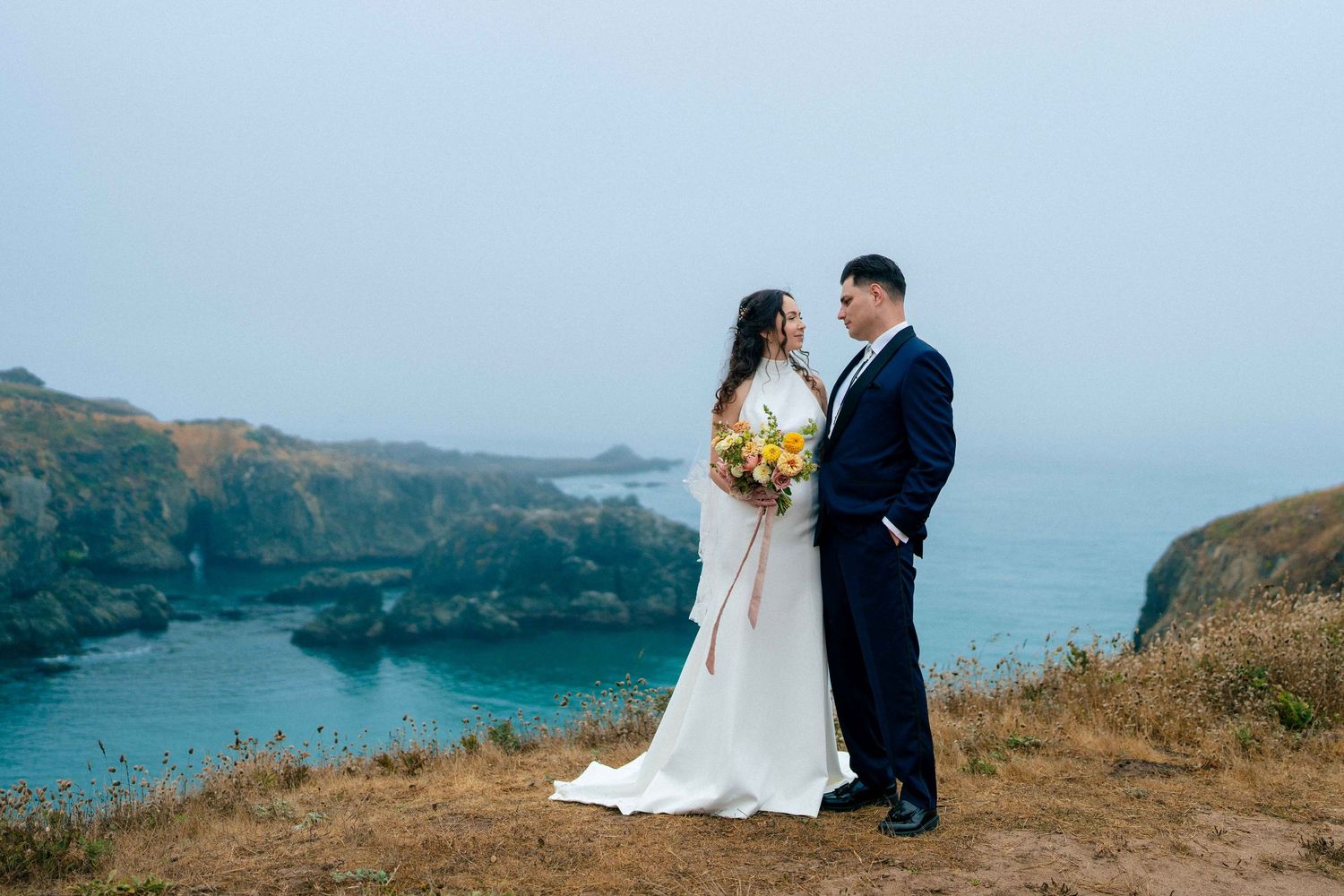 Couple embraces on dramatic coastal cliffside with rugged ocean coves and misty blue waters in the background.