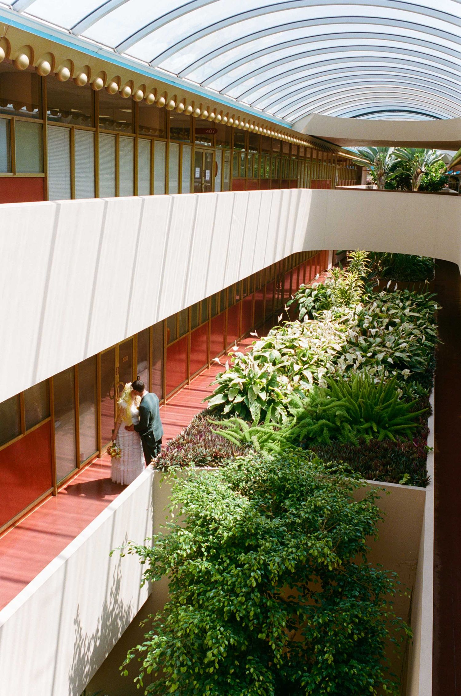 Interior atrium of a modern building featuring curved white walkways, lush green plants, and a glass ceiling with natural light.