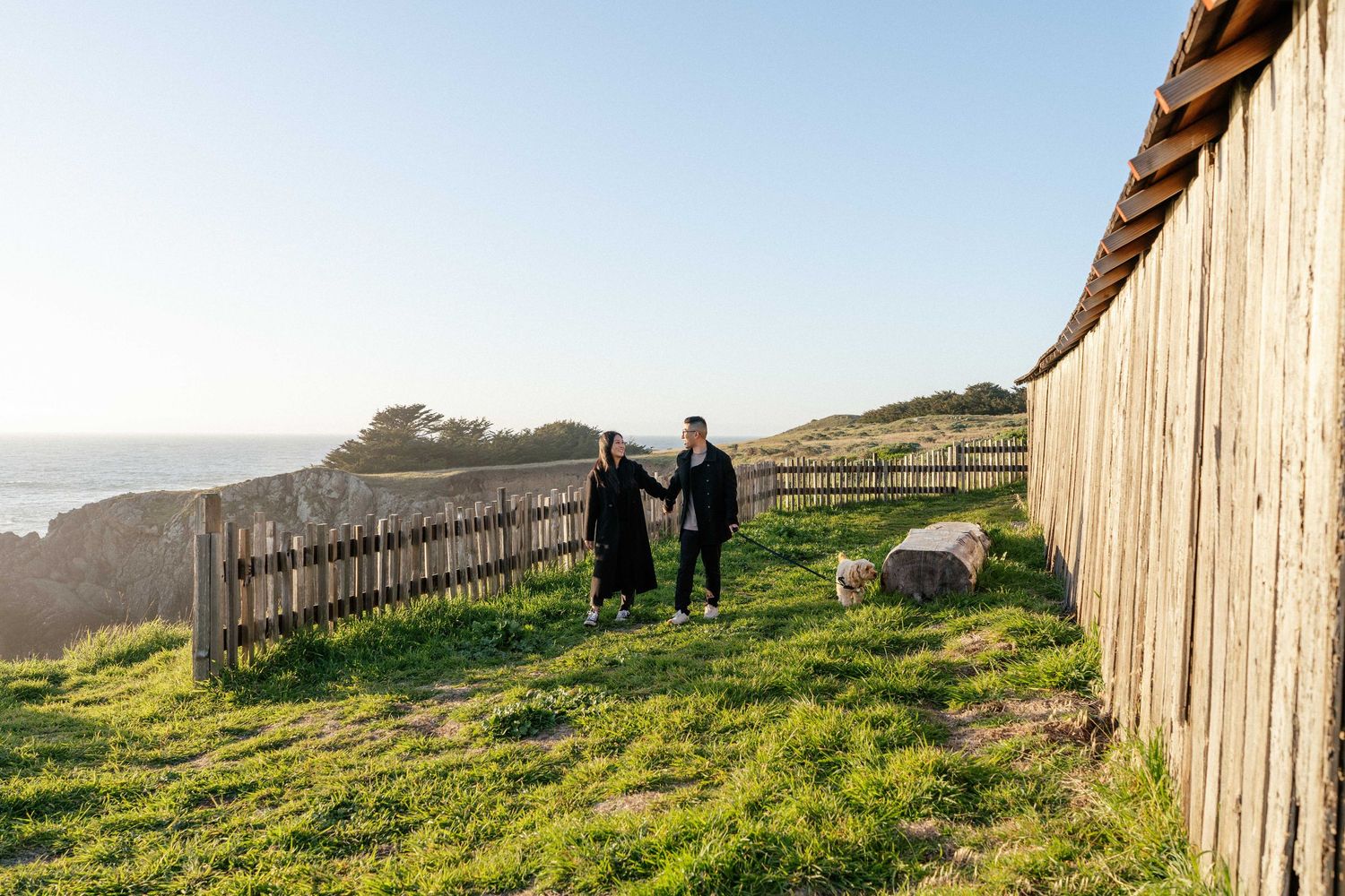 Couple holding hands walking on green grass along wooden fence with ocean view on sunny day.