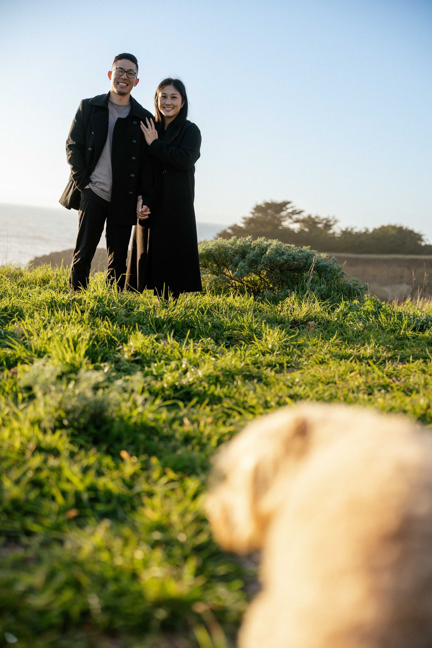 Couple smiling outdoors on green grass with a golden dog in foreground and coastal scenery behind them.