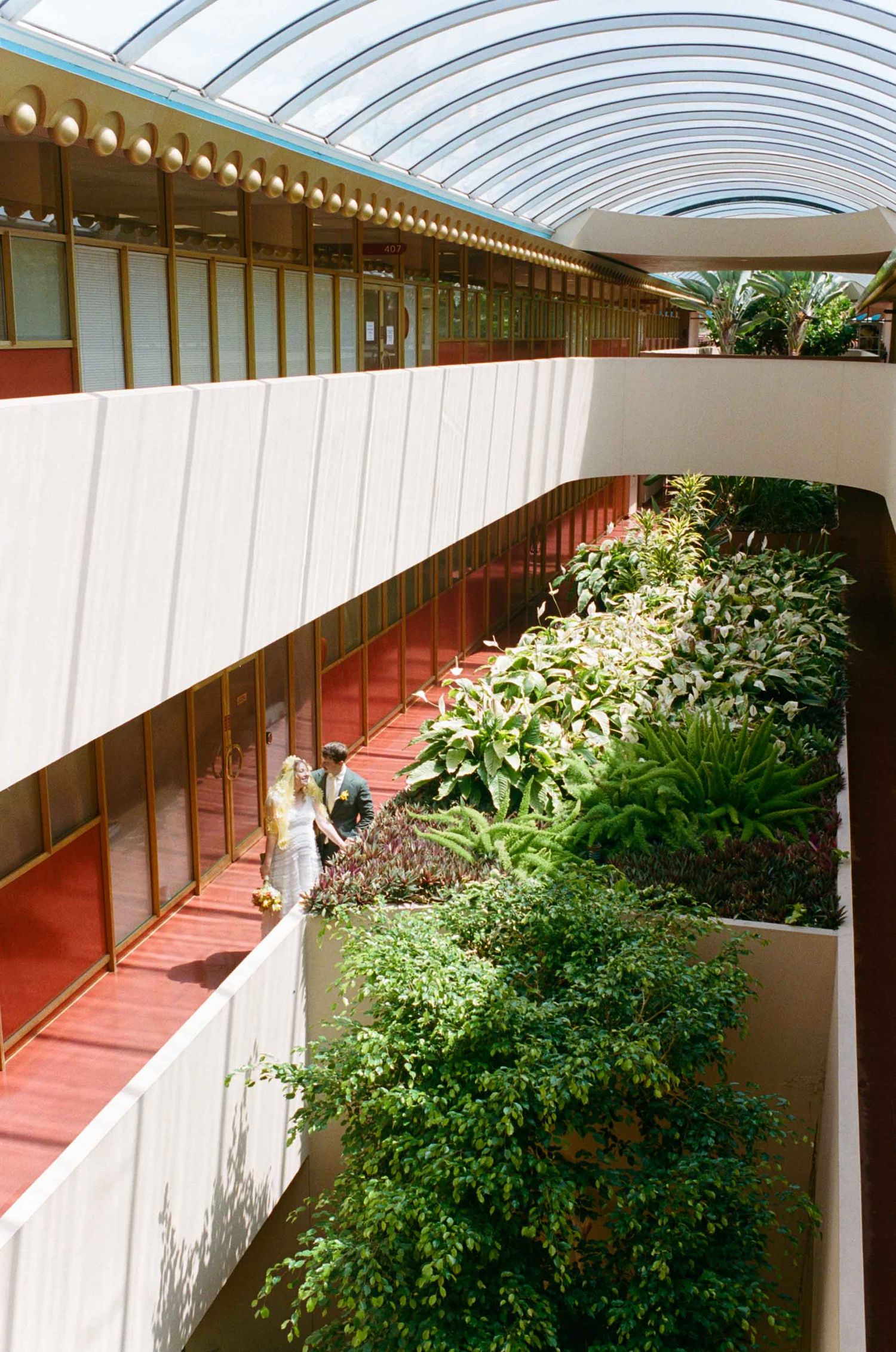 Interior atrium with curved glass ceiling, lush tropical plants, red walkways, and two people strolling through the garden corridor.