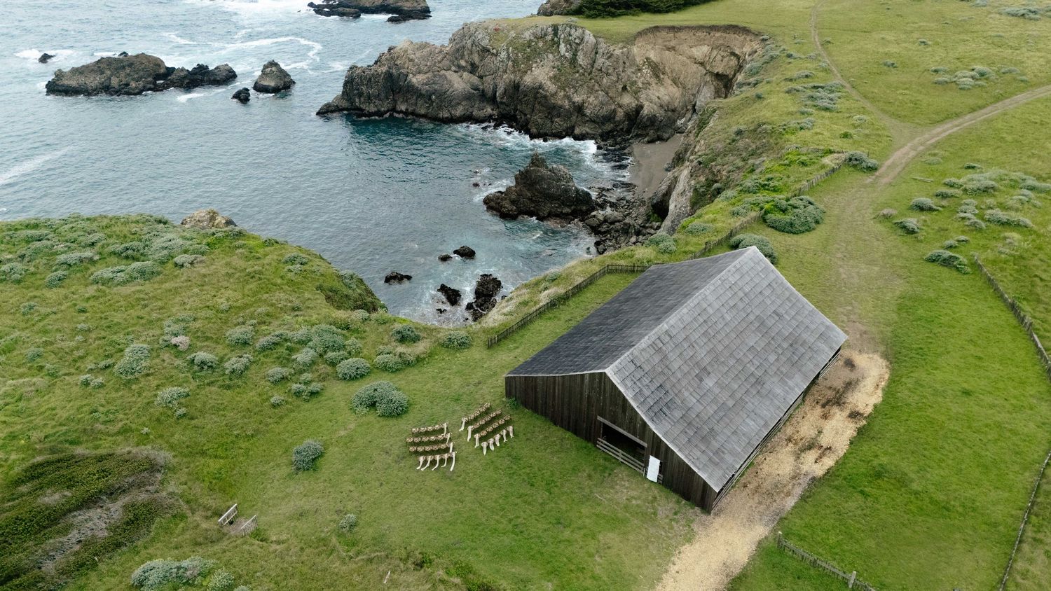 Aerial view of a rustic barn on green coastal cliffs overlooking rocky Pacific Ocean shoreline.