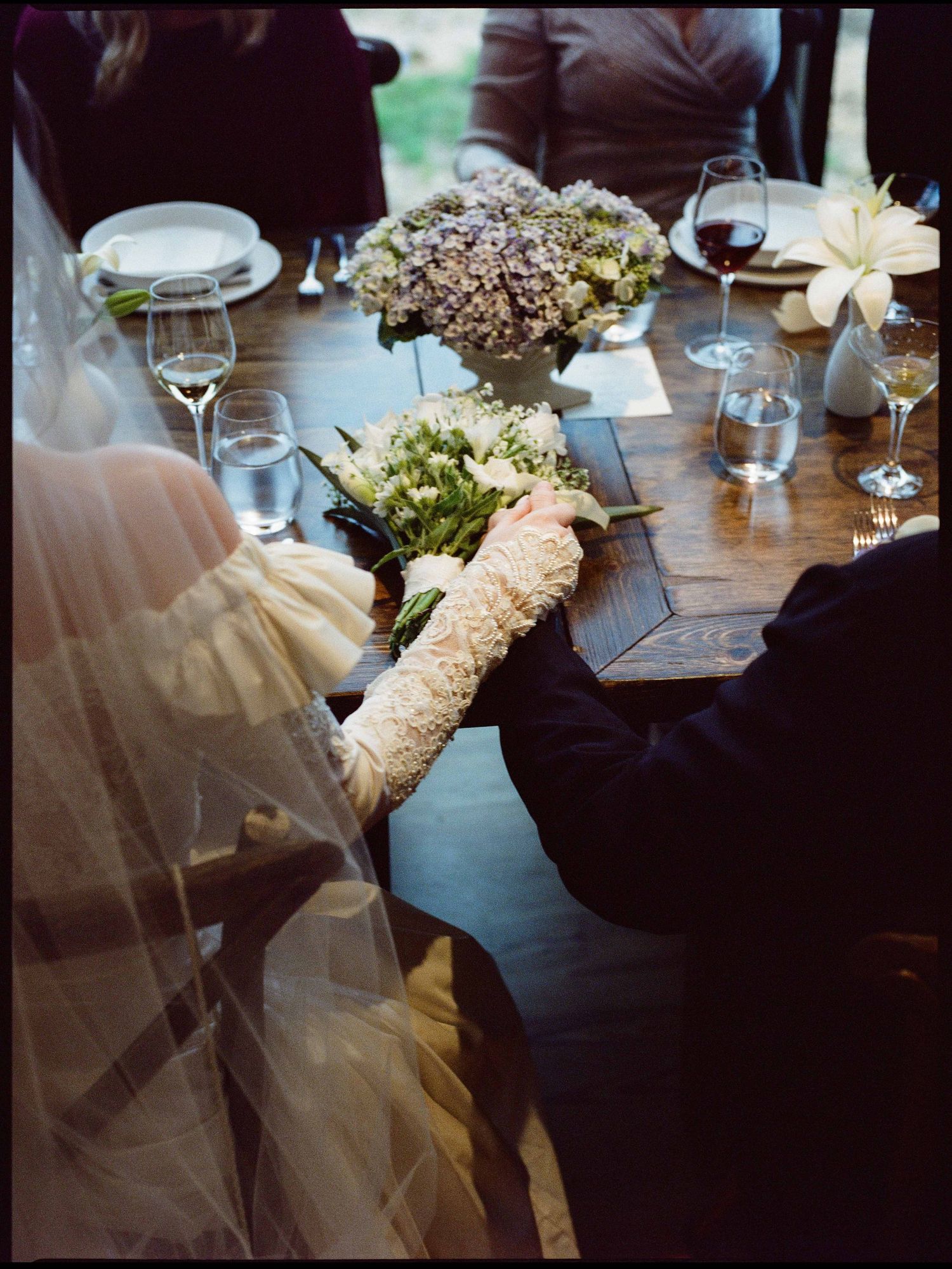 Bride in lace gloves holding bouquet at rustic wooden wedding reception table.