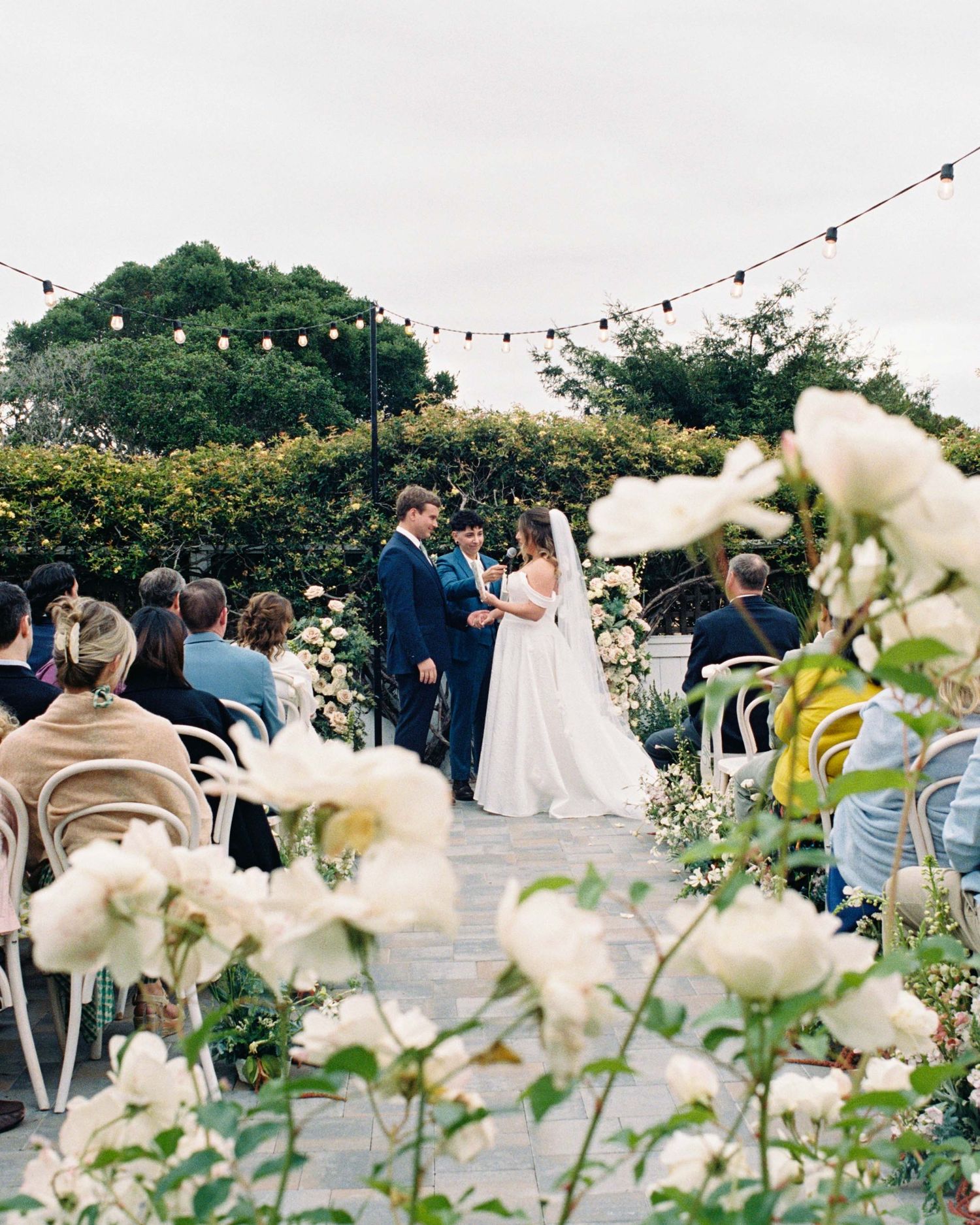 Outdoor wedding ceremony with couple at floral altar, string lights overhead, white roses in foreground, guests seated on patio.