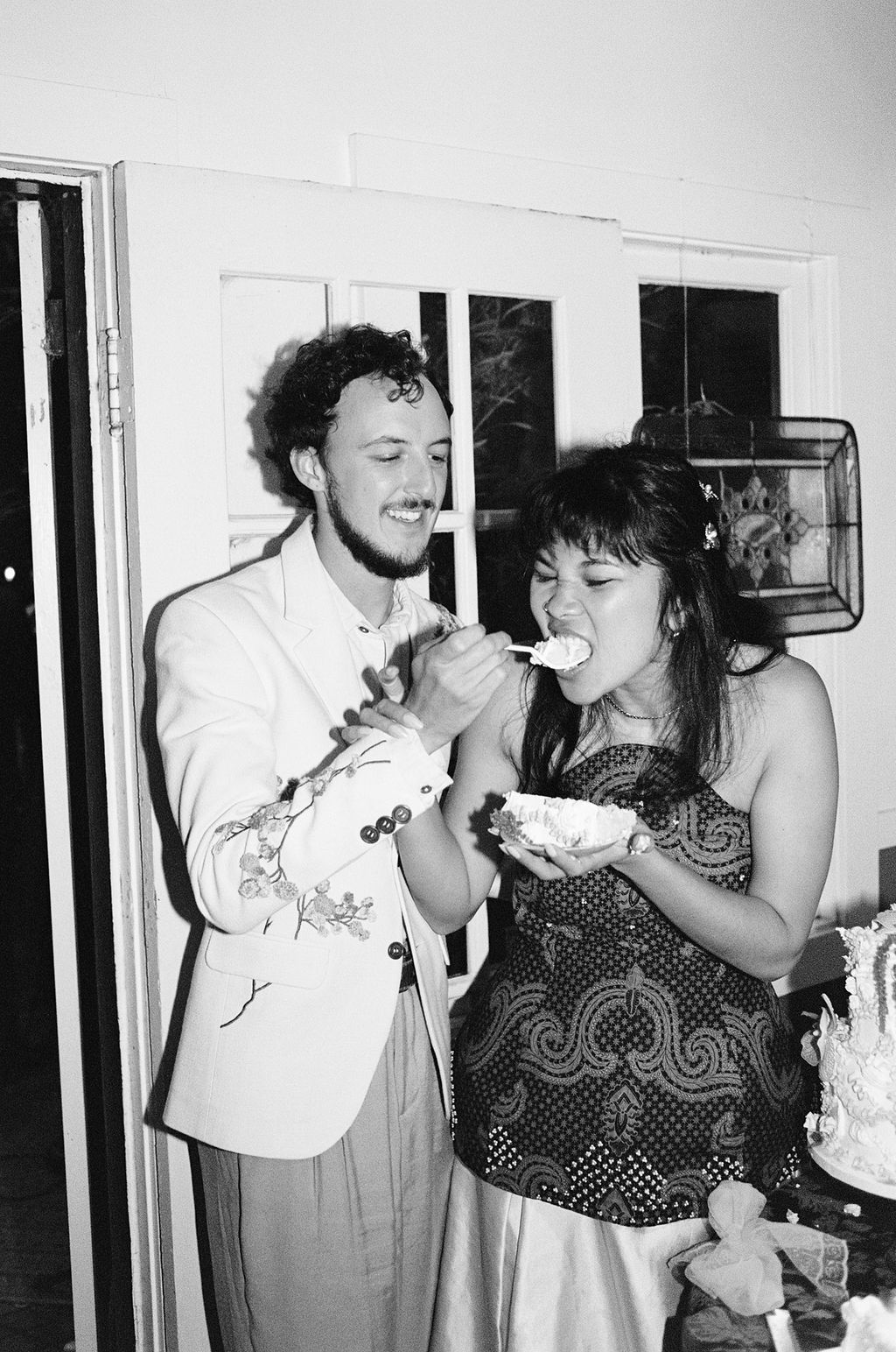 Black and white photo of a couple sharing wedding cake, smiling joyfully at an indoor celebration.