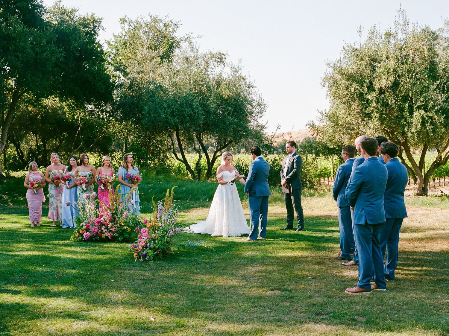 Wedding ceremony in a lush garden with bridesmaids in colorful dresses and groomsmen in blue suits.
