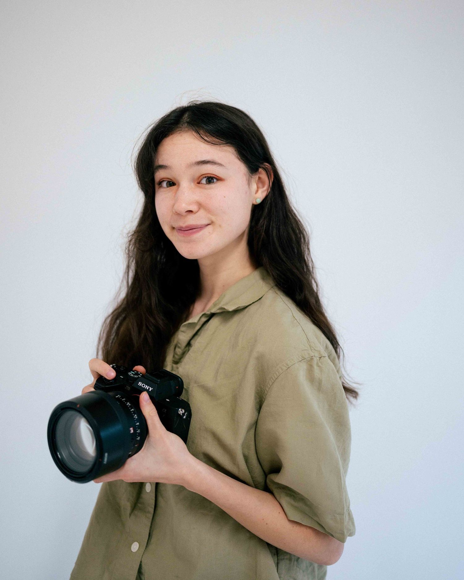 Photographer in olive green shirt holds professional camera against white background.