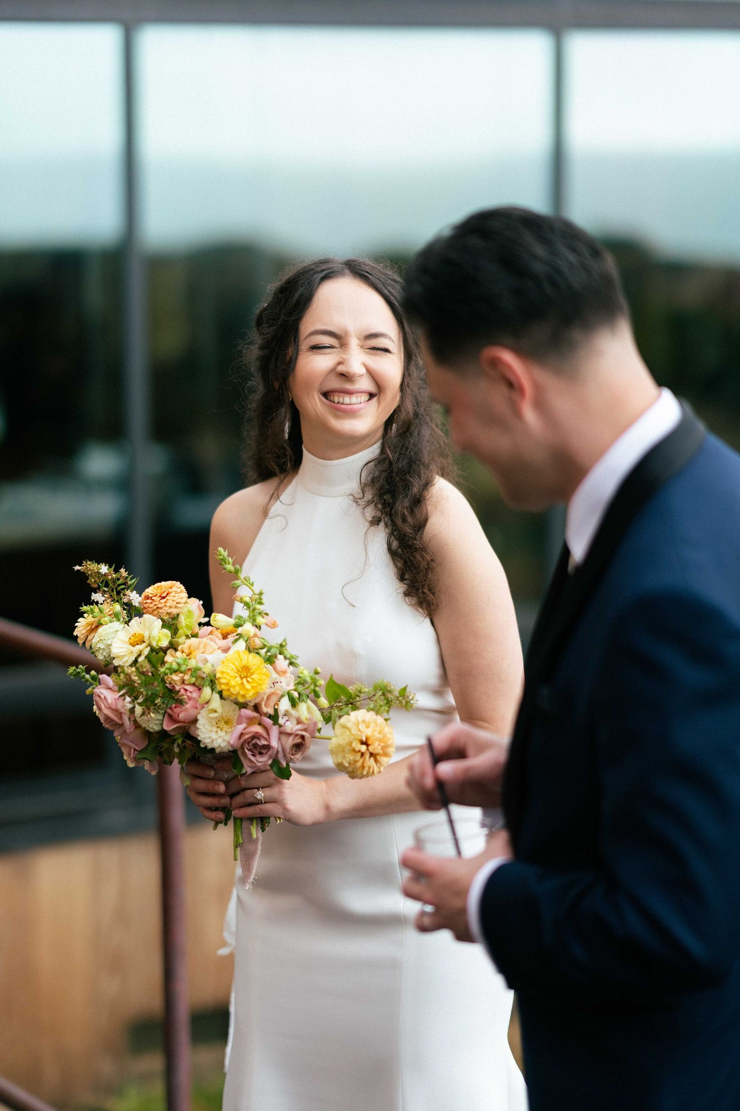 Smiling bride in white dress holding colorful bouquet laughing with groom in navy suit at outdoor wedding venue.