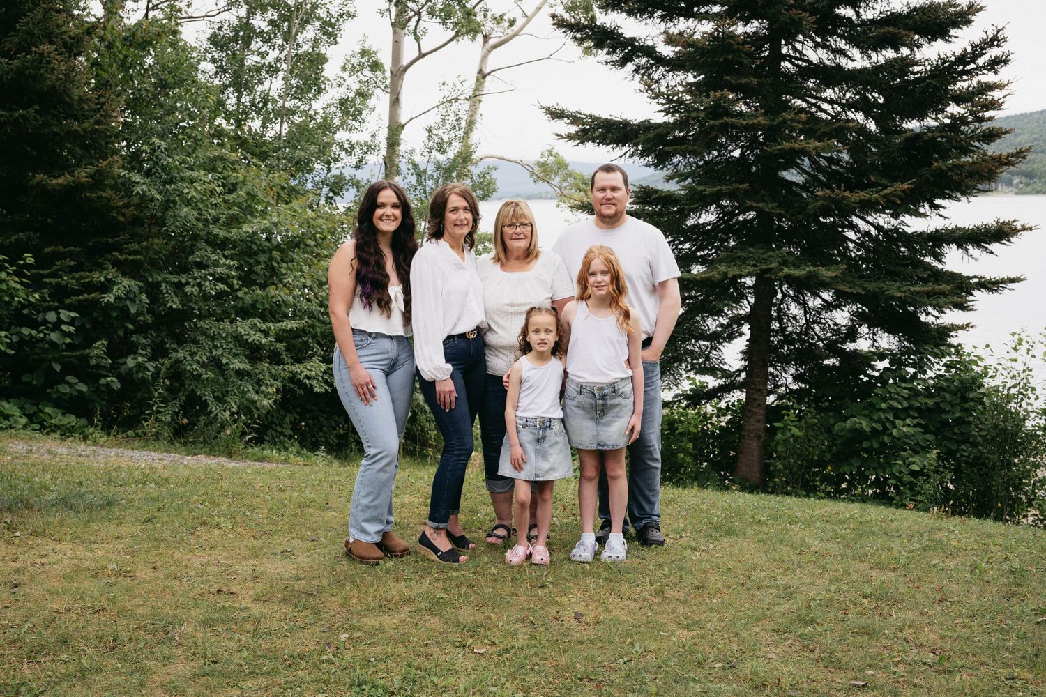 Family of six posing outdoors on green grass surrounded by tall trees in a casual white outfit photo session.