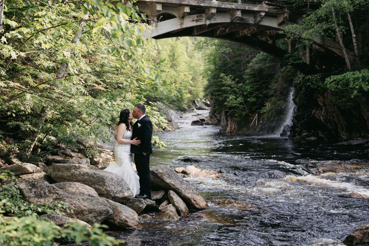 Bride and groom kissing on rocks beside a flowing river with a stone bridge and waterfall in a lush forest setting.