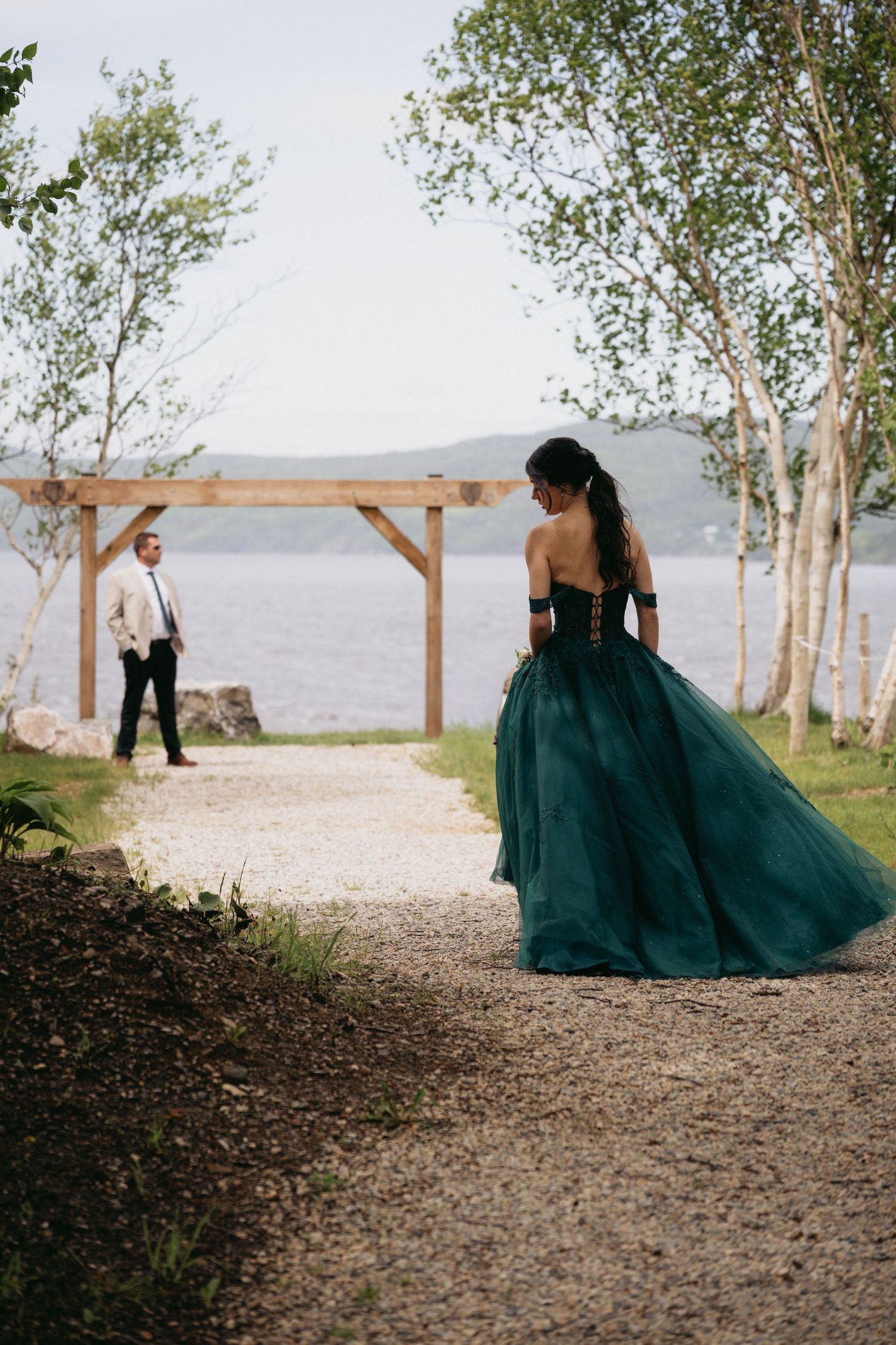 Bride in emerald green ball gown faces groom in white suit near wooden arbor by lakeside.