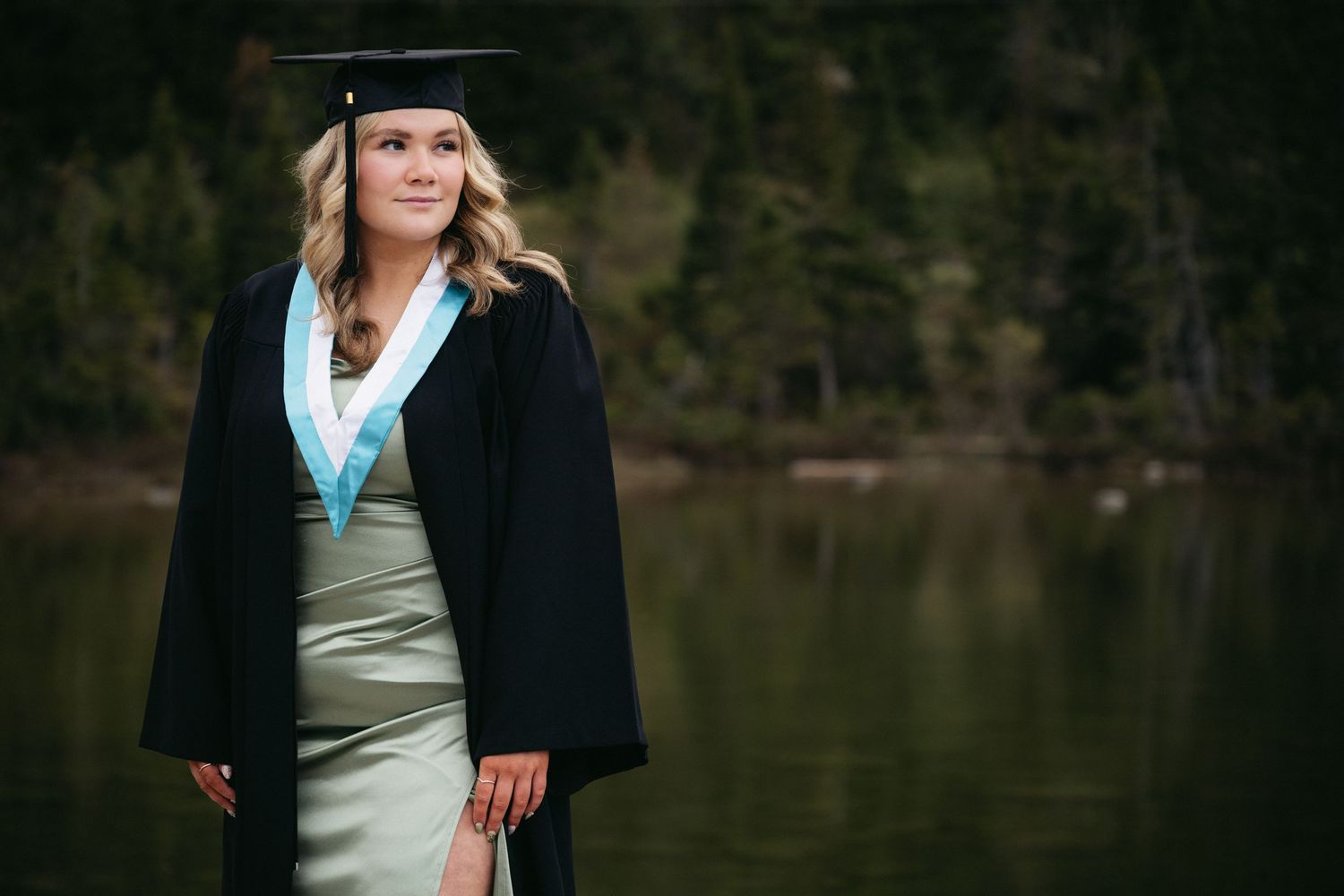 Young woman in graduation cap and gown posing outdoors near a lake with forest backdrop.