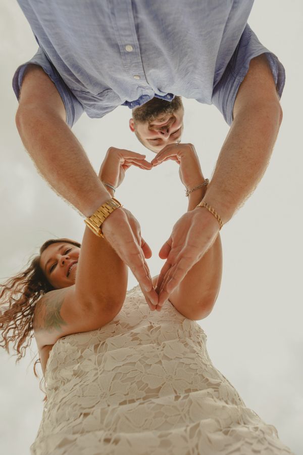 Couple making a heart shape with their hands, woman in white lace dress smiling, man in blue shirt, gold watches.