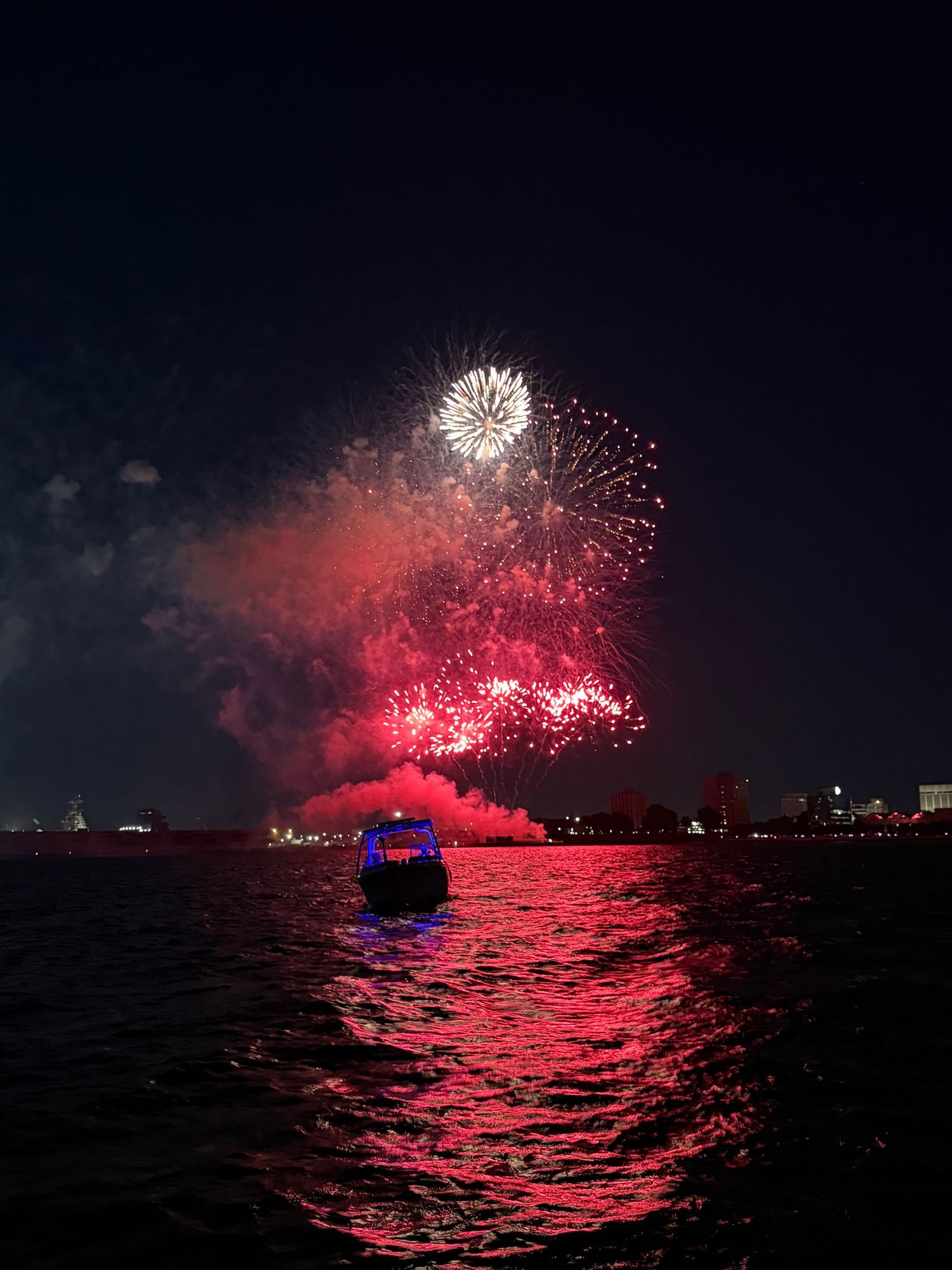 Red fireworks illuminate the night sky over water with a boat silhouetted in the foreground during a festive display.