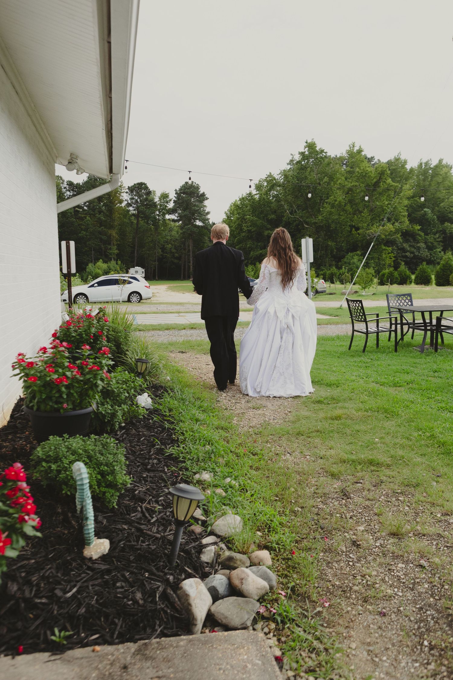 Bride and groom walking hand in hand along a garden path beside a white building on their wedding day.