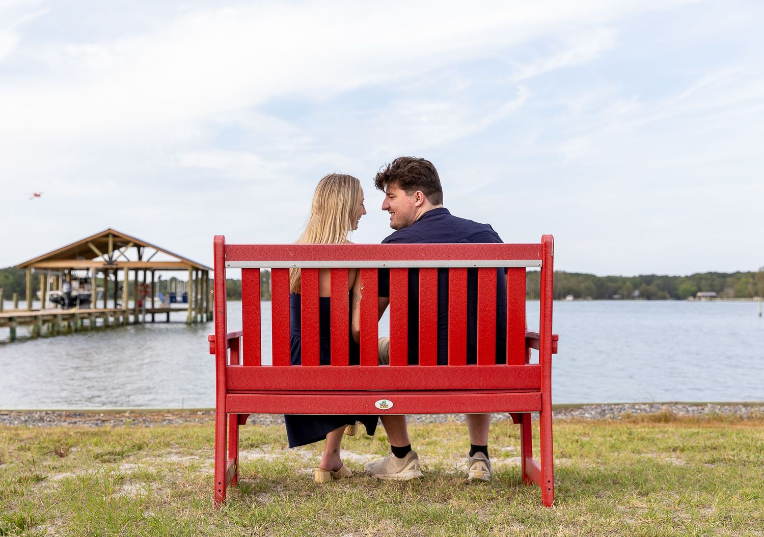 Couple sitting together on a red bench facing a waterfront dock and boathouse.