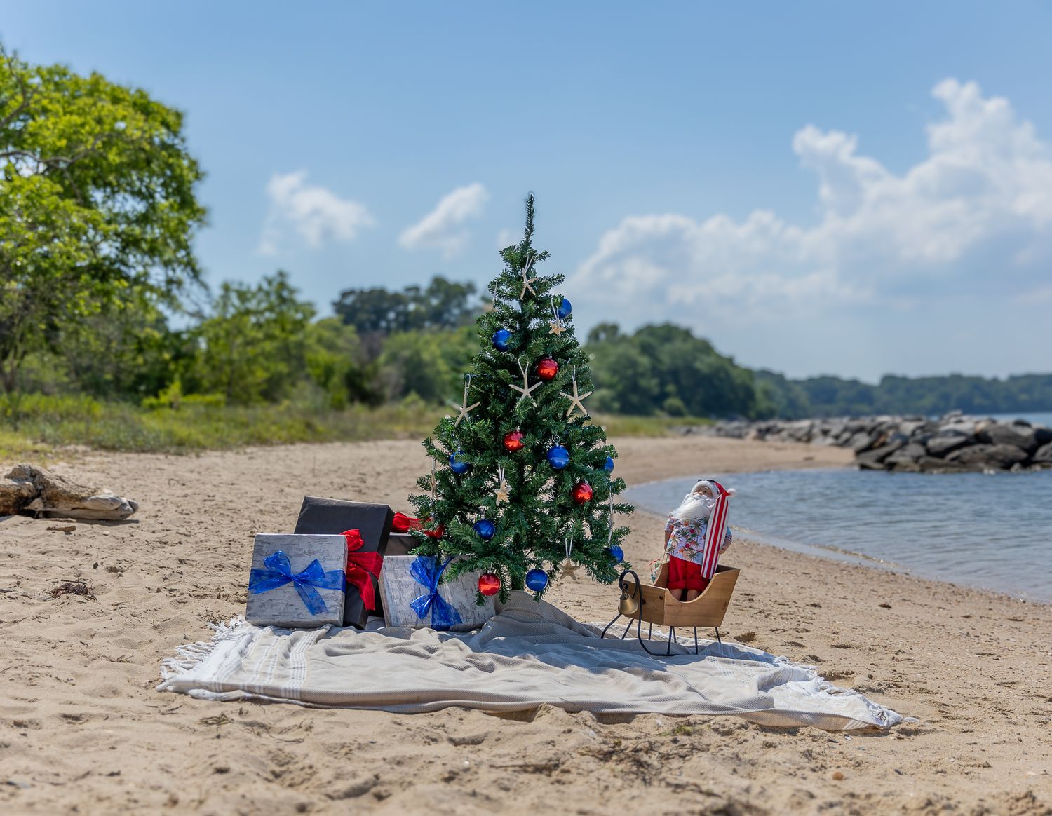 A decorated Christmas tree with gifts and a sleigh on a sandy beach with blue skies and trees in the background.