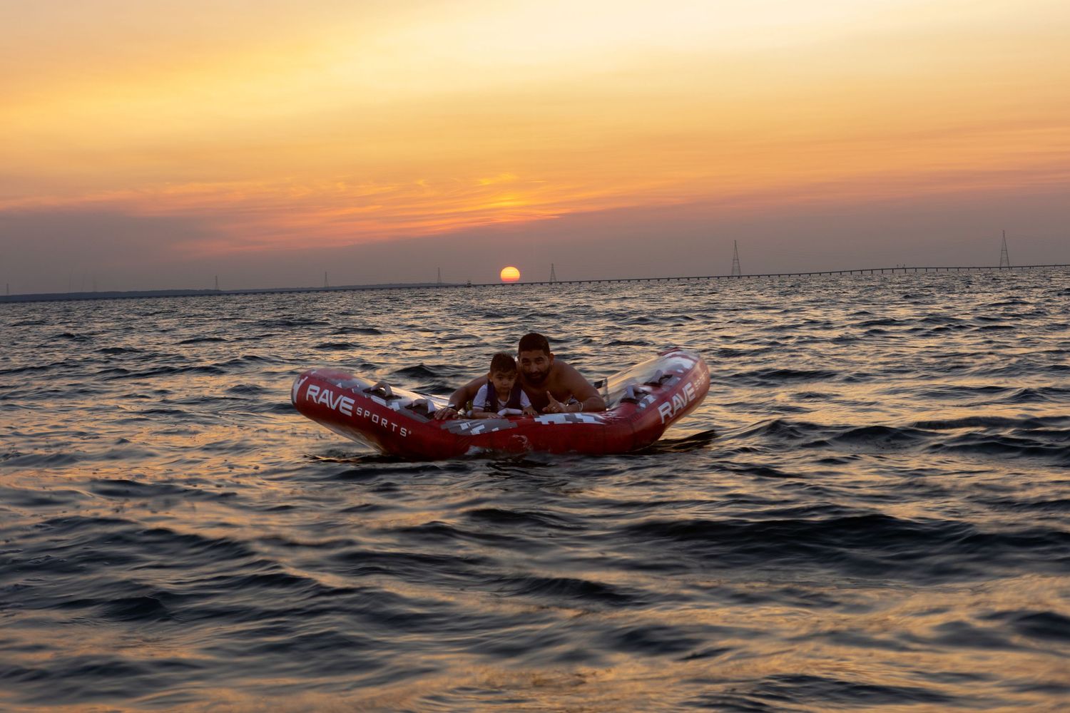 Person floating on inflatable raft in ocean during vibrant golden sunset with distant bridge on horizon.