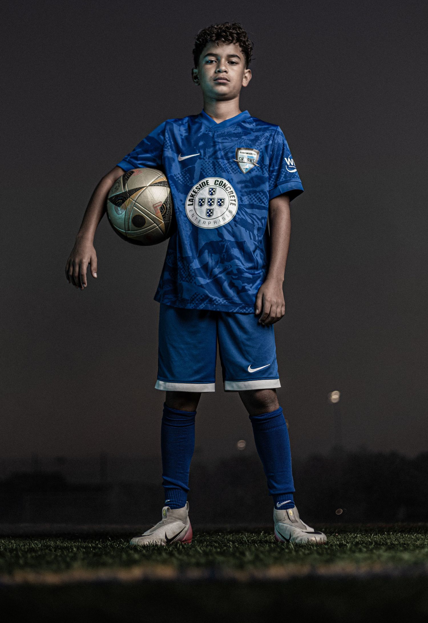 Young soccer player in blue Nike uniform holds a ball on a grass field during a dramatic nighttime portrait shoot.