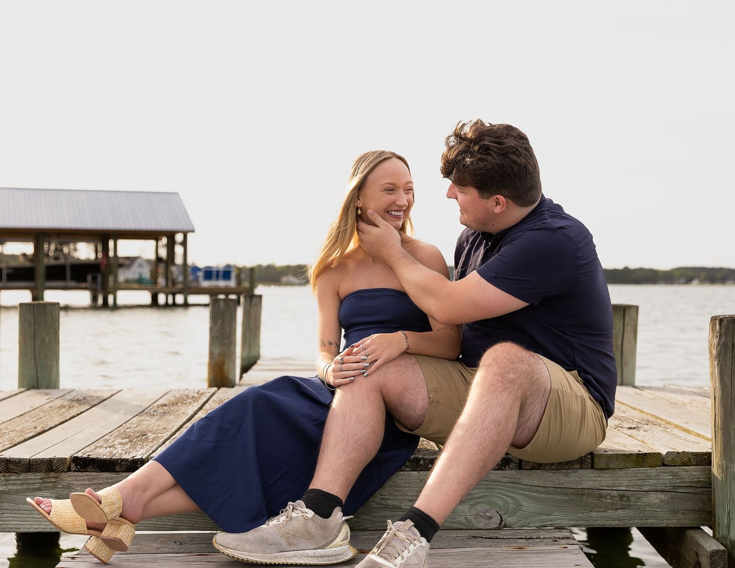 Couple sitting on a wooden dock by the water, sharing a romantic moment with a pier in the background.