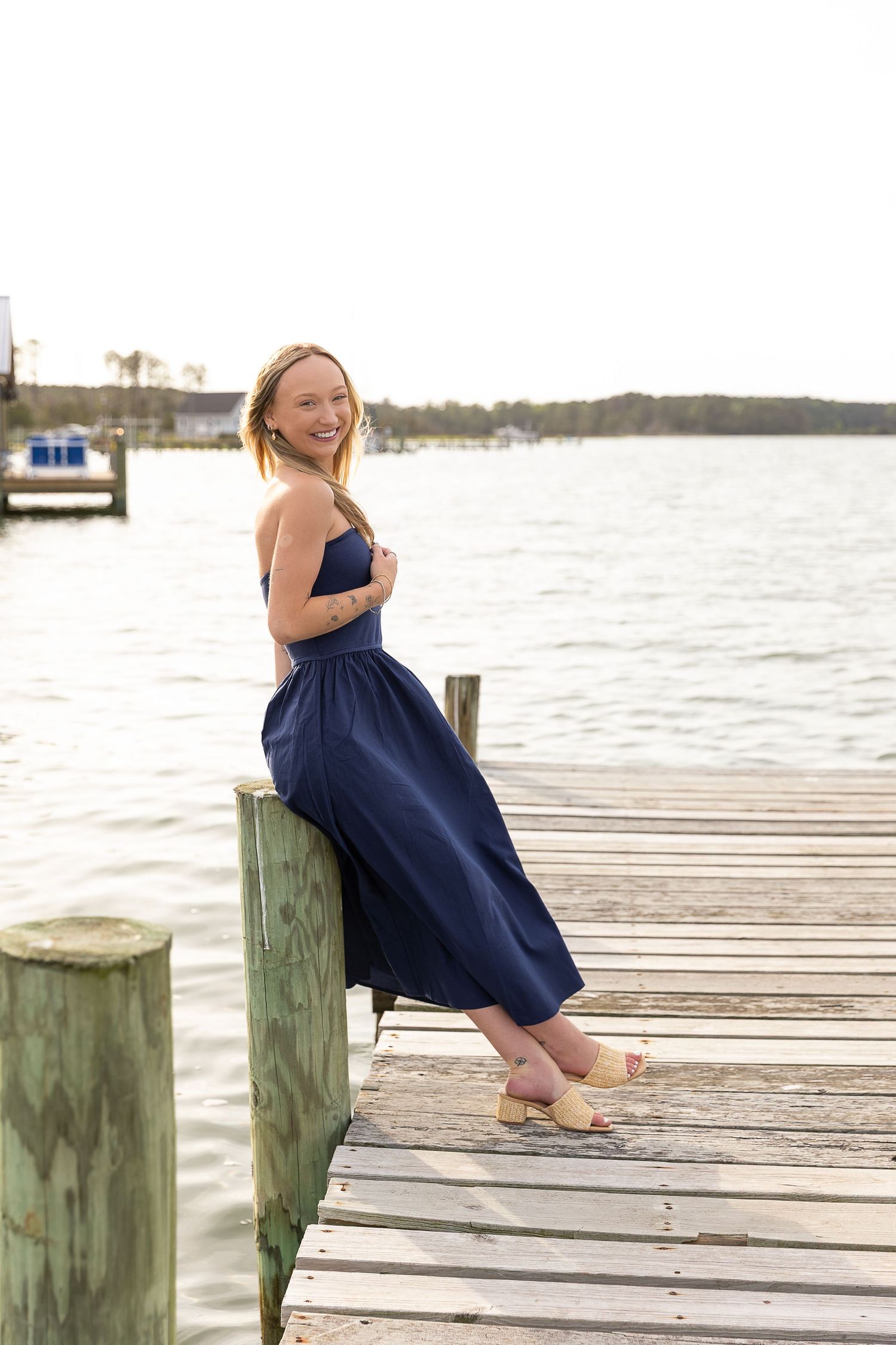 Young woman in navy blue strapless dress smiling on wooden dock by water.