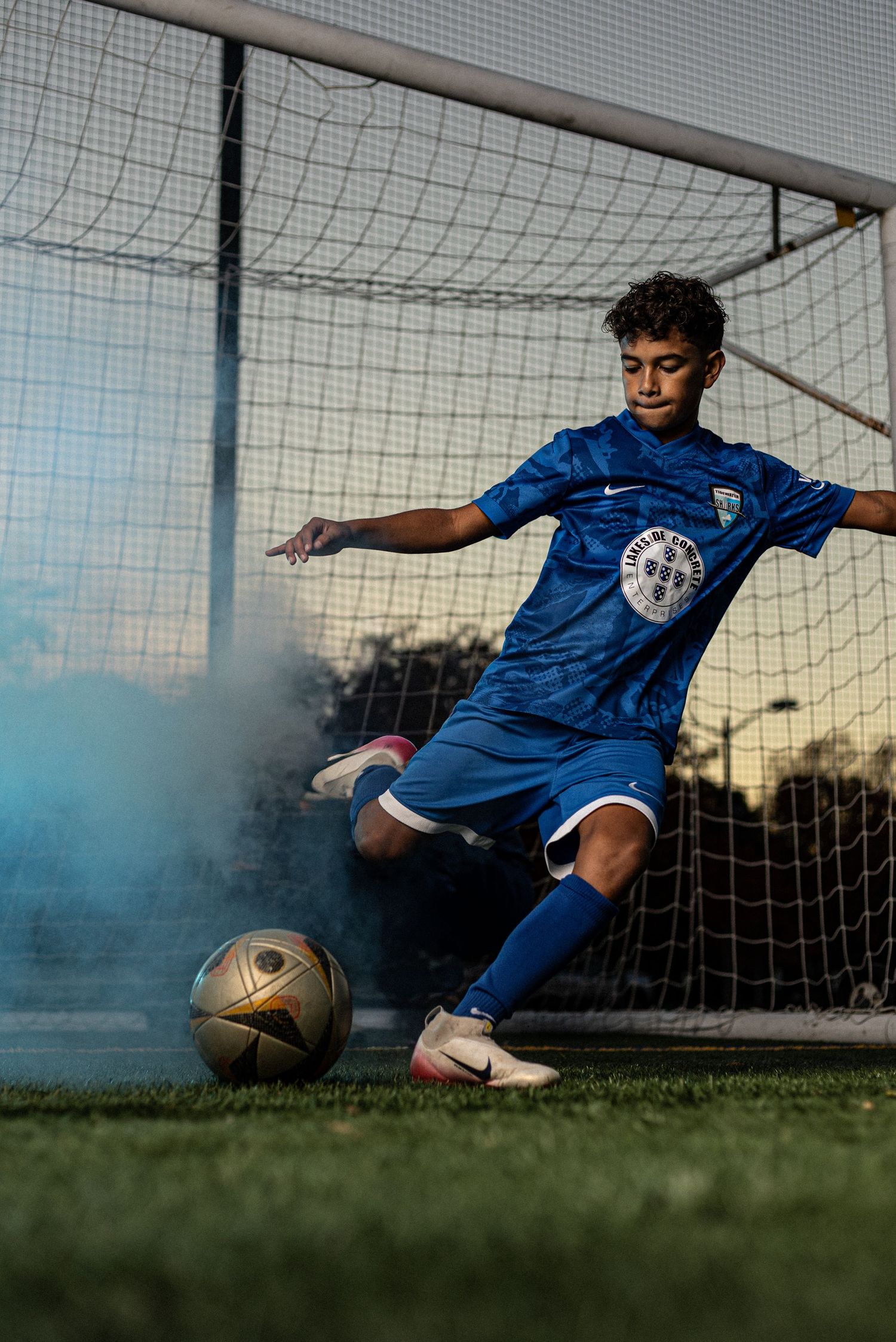 Young soccer player in blue Nike kit kicking a ball near a goal with blue smoke on a grass field.