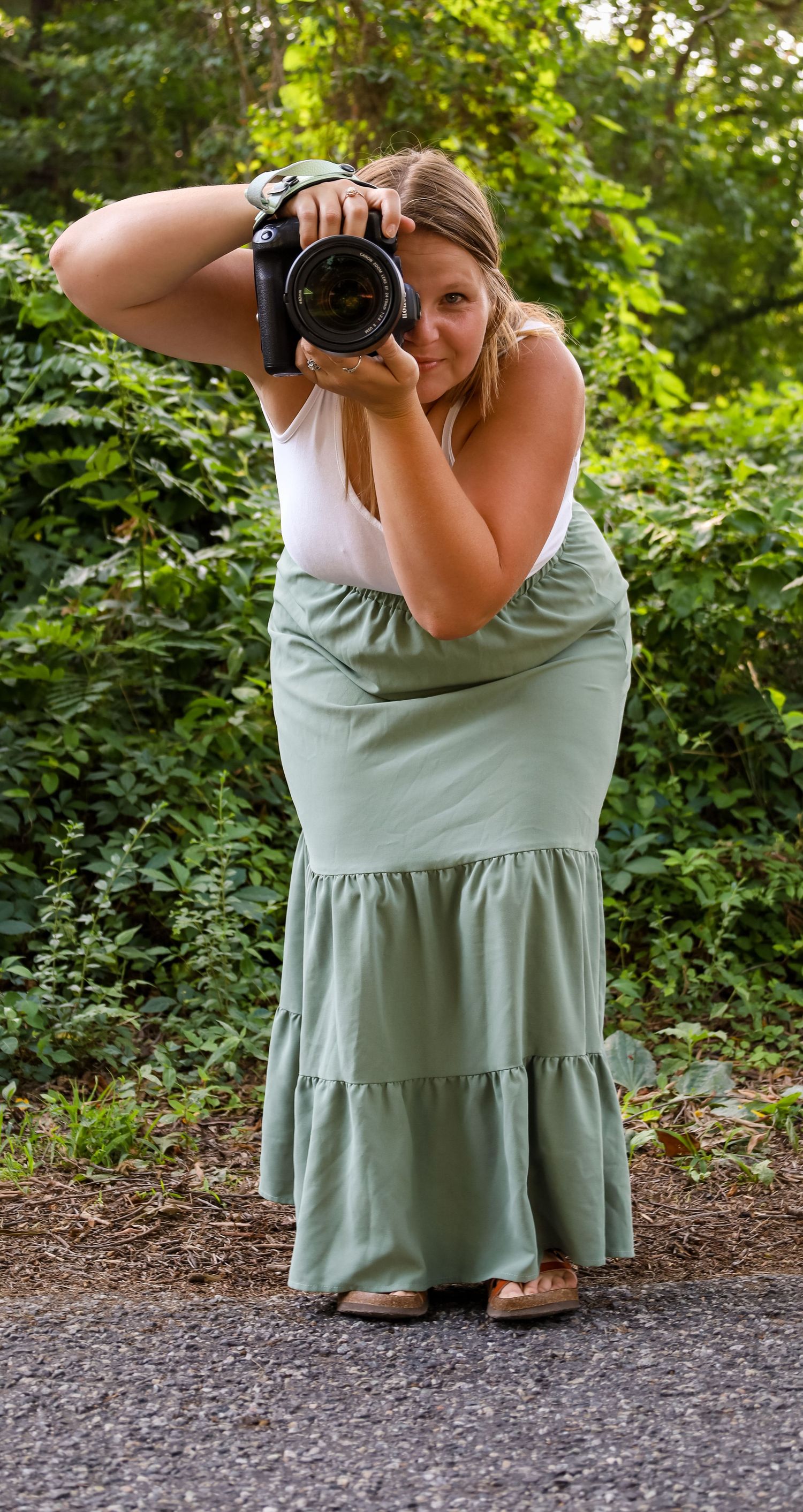 Woman in green tiered maxi skirt holds DSLR camera up to her face outdoors surrounded by lush greenery.