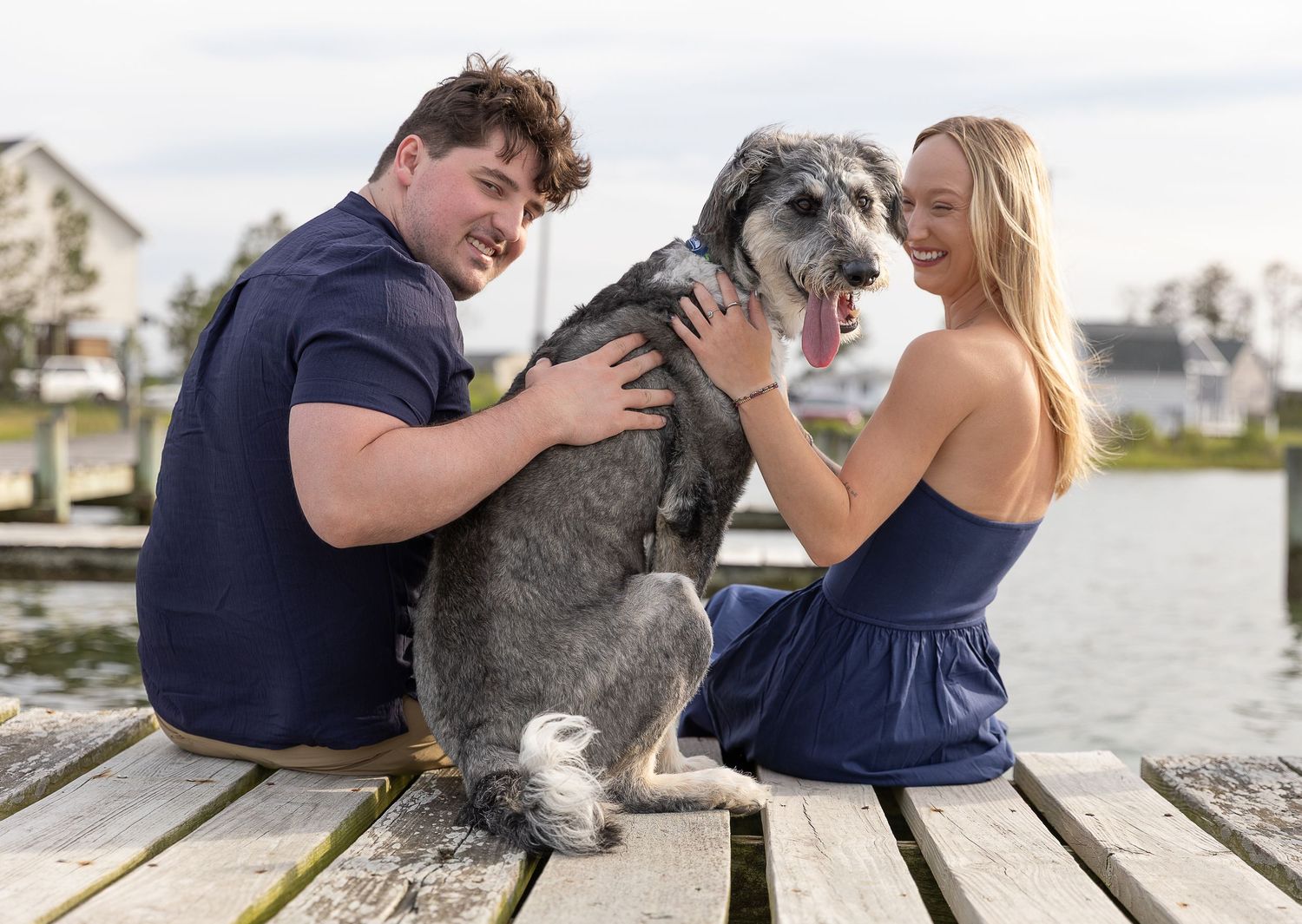 A happy couple sits on a wooden dock petting their fluffy gray dog near a waterfront.