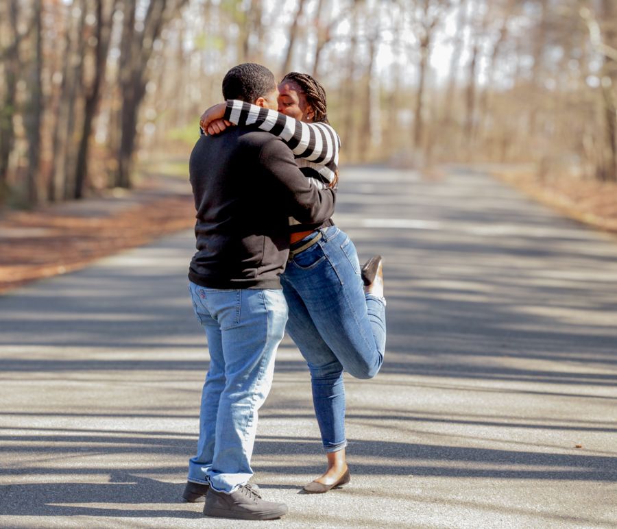 Couple sharing a romantic embrace on a tree-lined road, with woman lifting her leg joyfully.