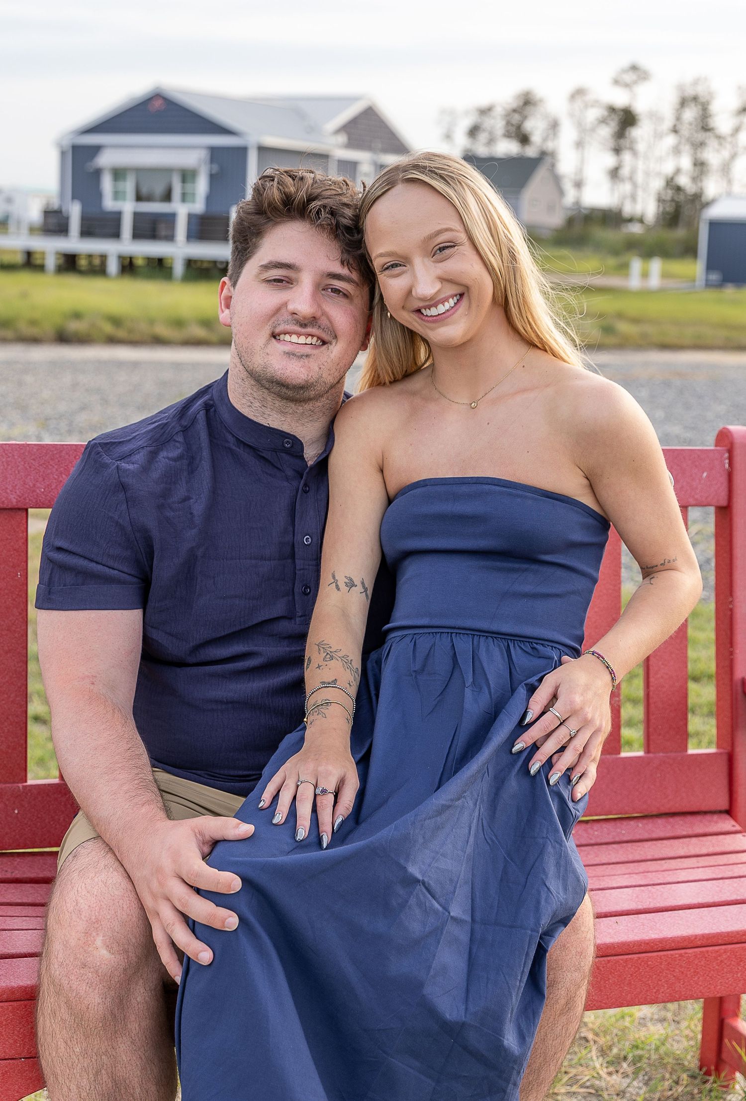 Smiling couple sitting on a red bench outdoors with a waterfront home in the background.