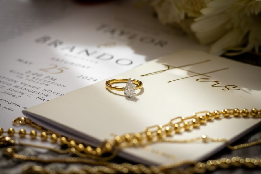 Gold diamond engagement ring resting on elegant wedding invitation with pearl necklace and white flowers in background.