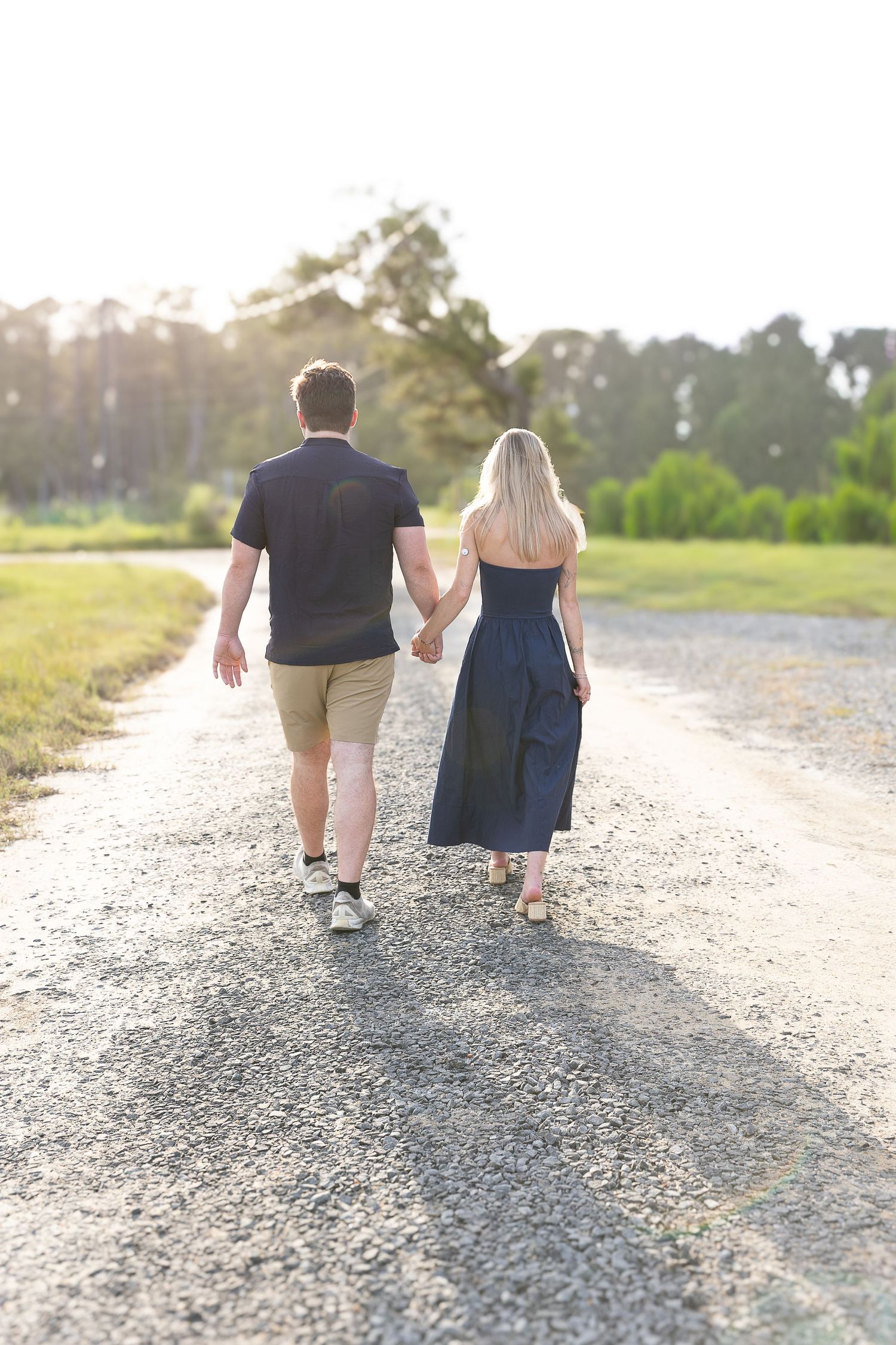 Couple holding hands walking down sunny gravel path surrounded by green trees.