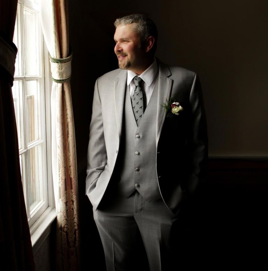A man in a gray three-piece suit with a floral boutonniere stands by a window, smiling in a dimly lit room.