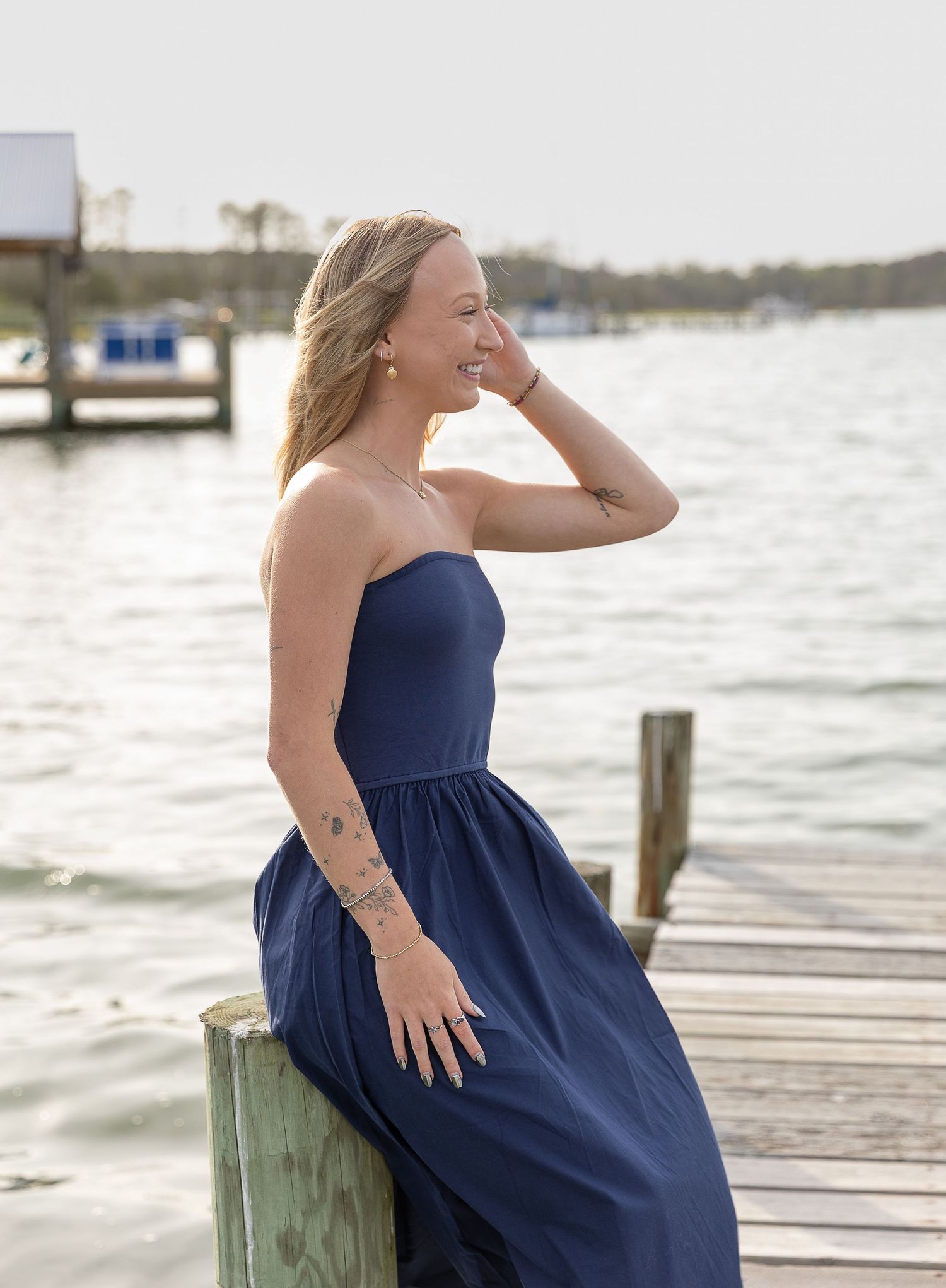 Young woman in strapless navy blue dress sitting on a dock, laughing with water in the background.