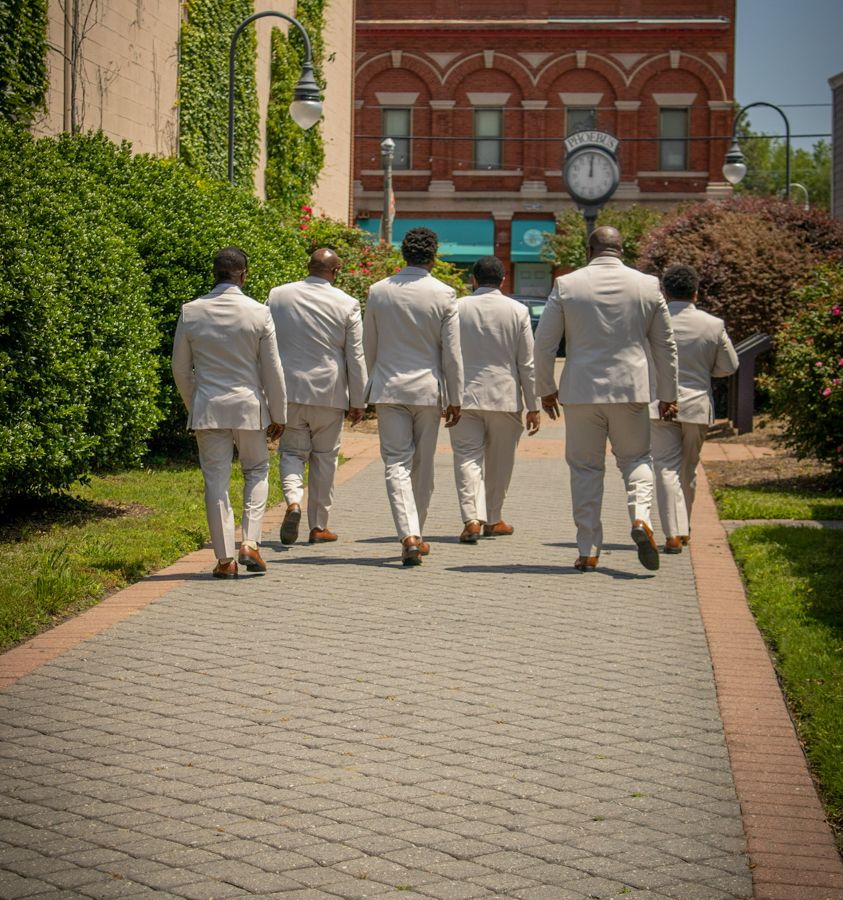 Six men in white suits walk away down a brick pathway past a red brick building and clock on a sunny day.