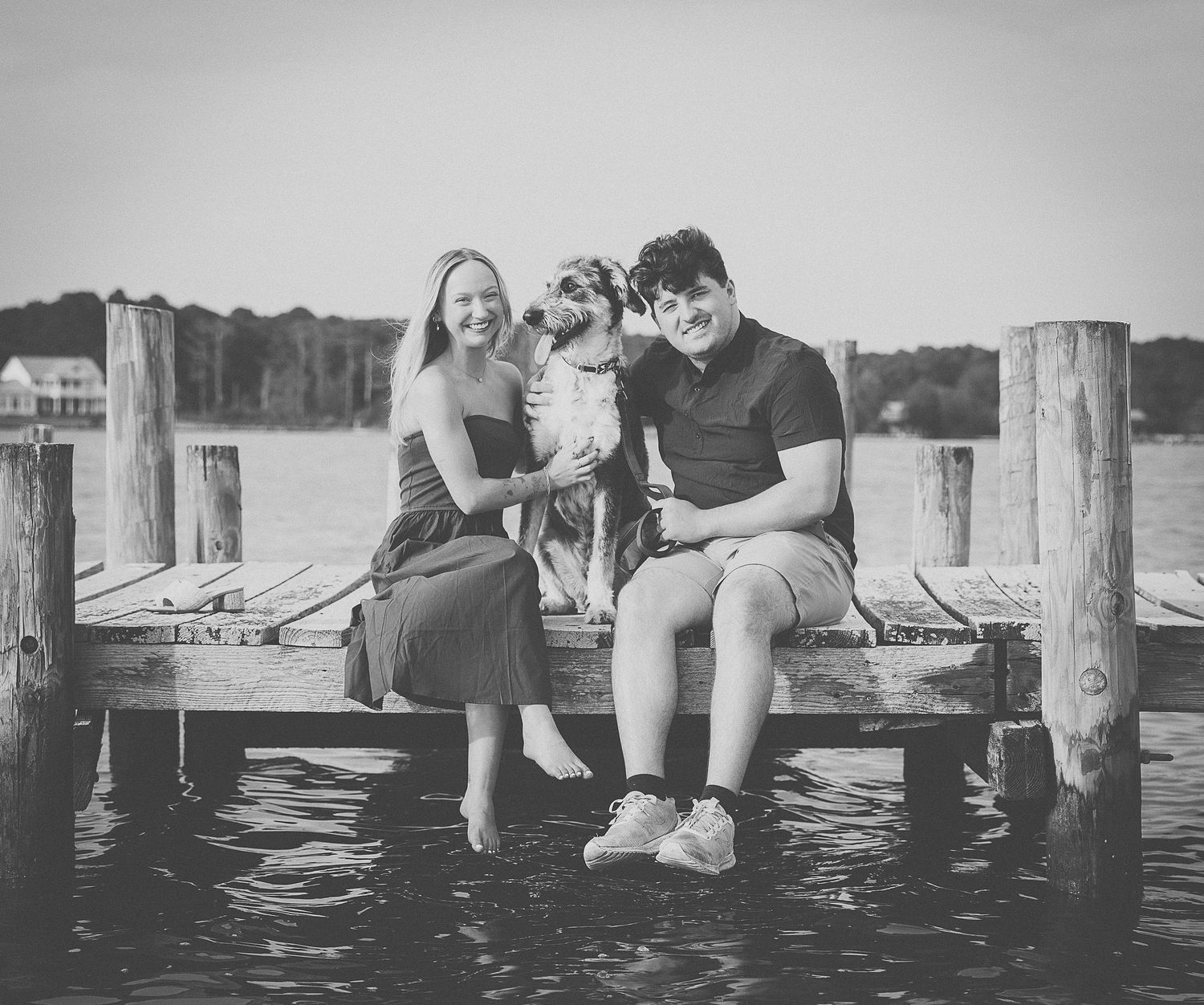 Couple sitting on a wooden dock with their dog by the water in a black and white photo.